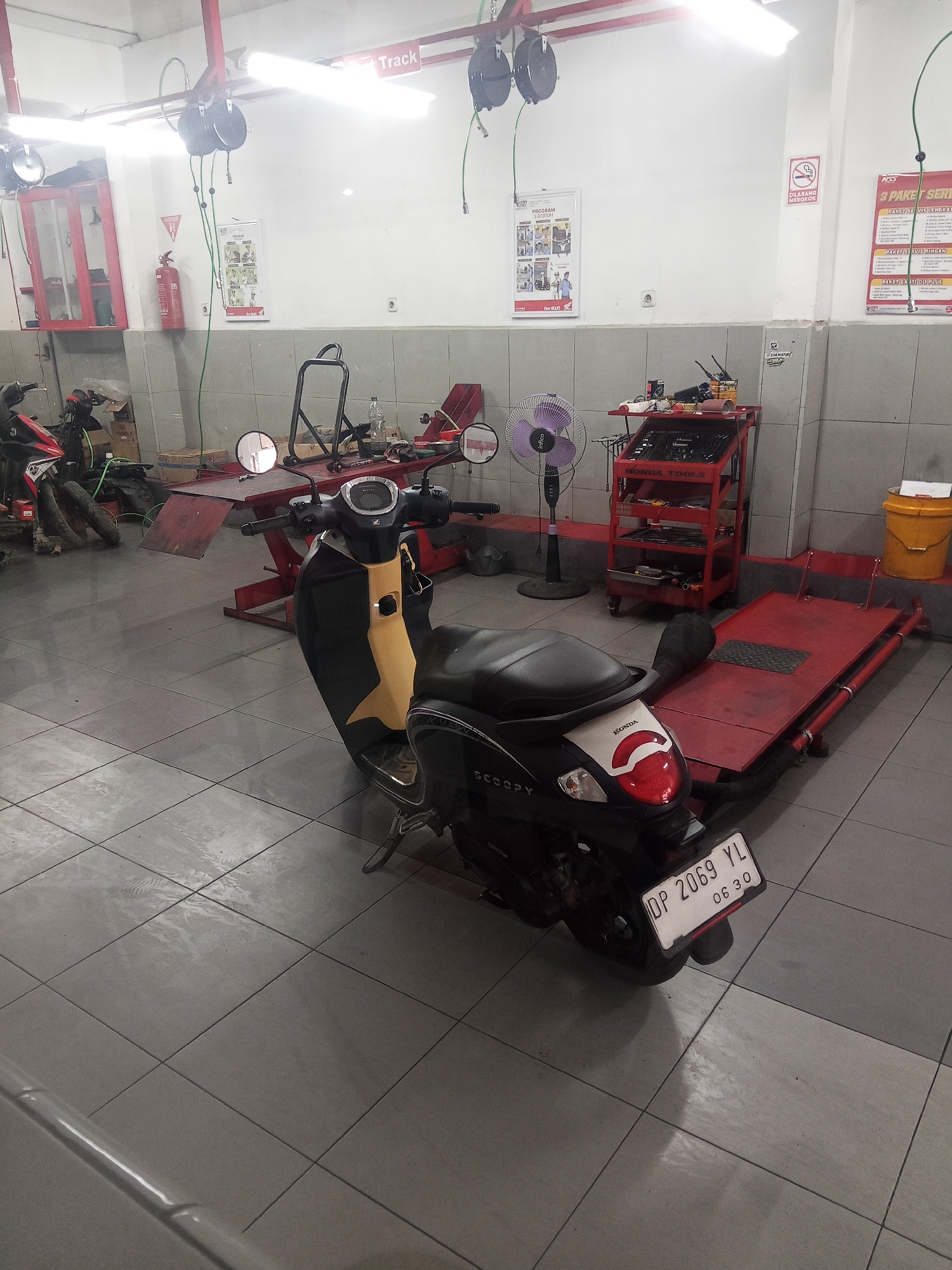 This is a photograph of a motorcycle repair shop in Rinding Batu, Indonesia. A black and yellow Honda Scoopy motorcycle is parked in the center of the frame, facing away from the viewer. It is positioned on the grey tiled floor. To the left of the motorcycle, there is a red hydraulic lift with a motorcycle on it. In the background, there is a red tool cart labeled "HONDA TOOLS", a purple oscillating fan, and a yellow bucket. Signs on the wall include "Track", "PROGRAM BEE SIN", and a "DILARANG MEROKOK" (No Smoking) sign. There is also a fire extinguisher mounted on the wall. The lighting appears to be fluorescent, typical of an indoor commercial space.