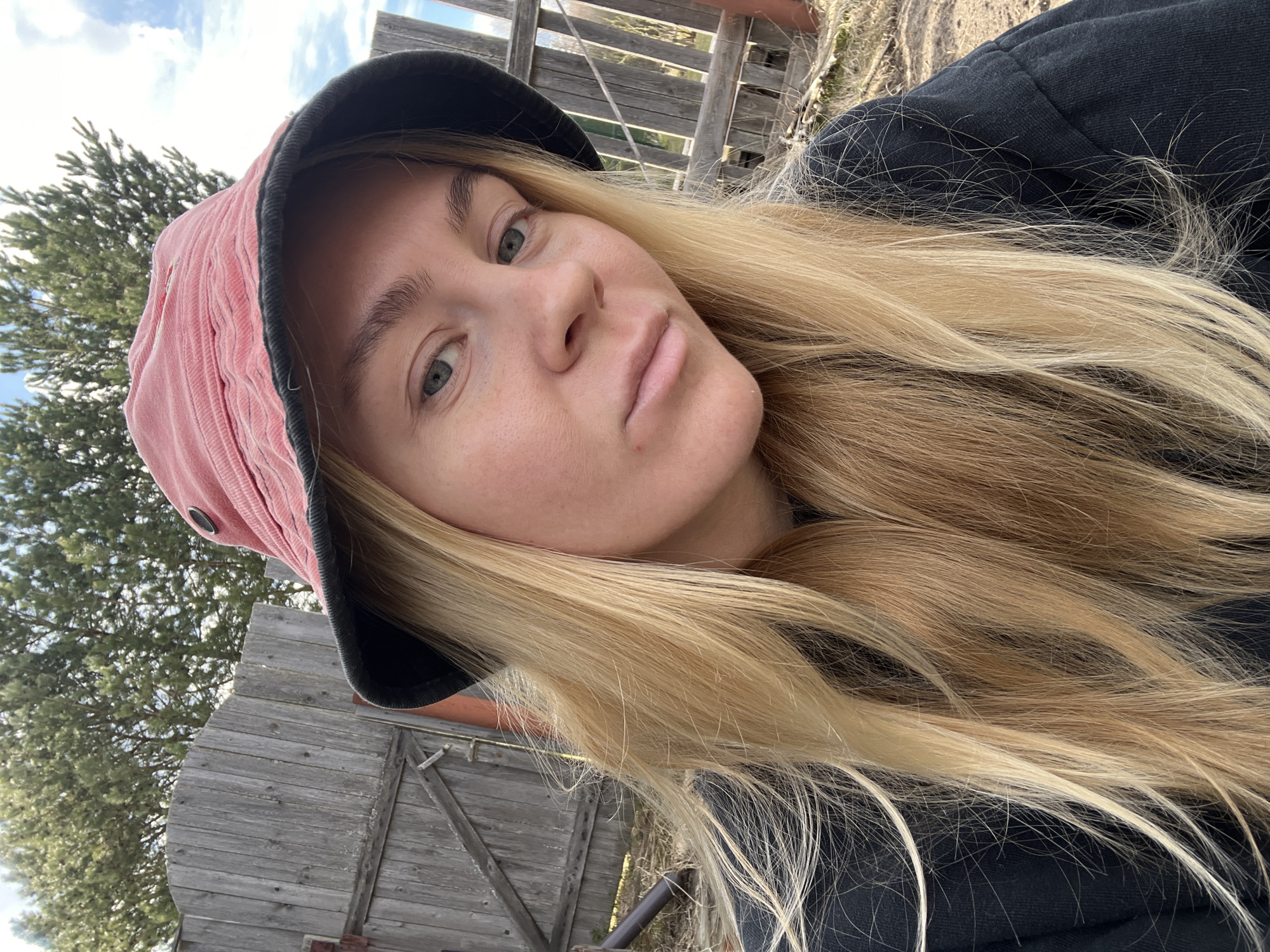 A selfie of a blonde woman wearing a pink corduroy bucket hat with a black brim. She has fair skin, green eyes, and full lips. Her hair is long and straight. She is wearing a black top or jacket. The background includes a weathered wooden fence and a tree. The sky is visible in the upper left corner. The location is likely near Chernysovo, Russia due to the prompt.