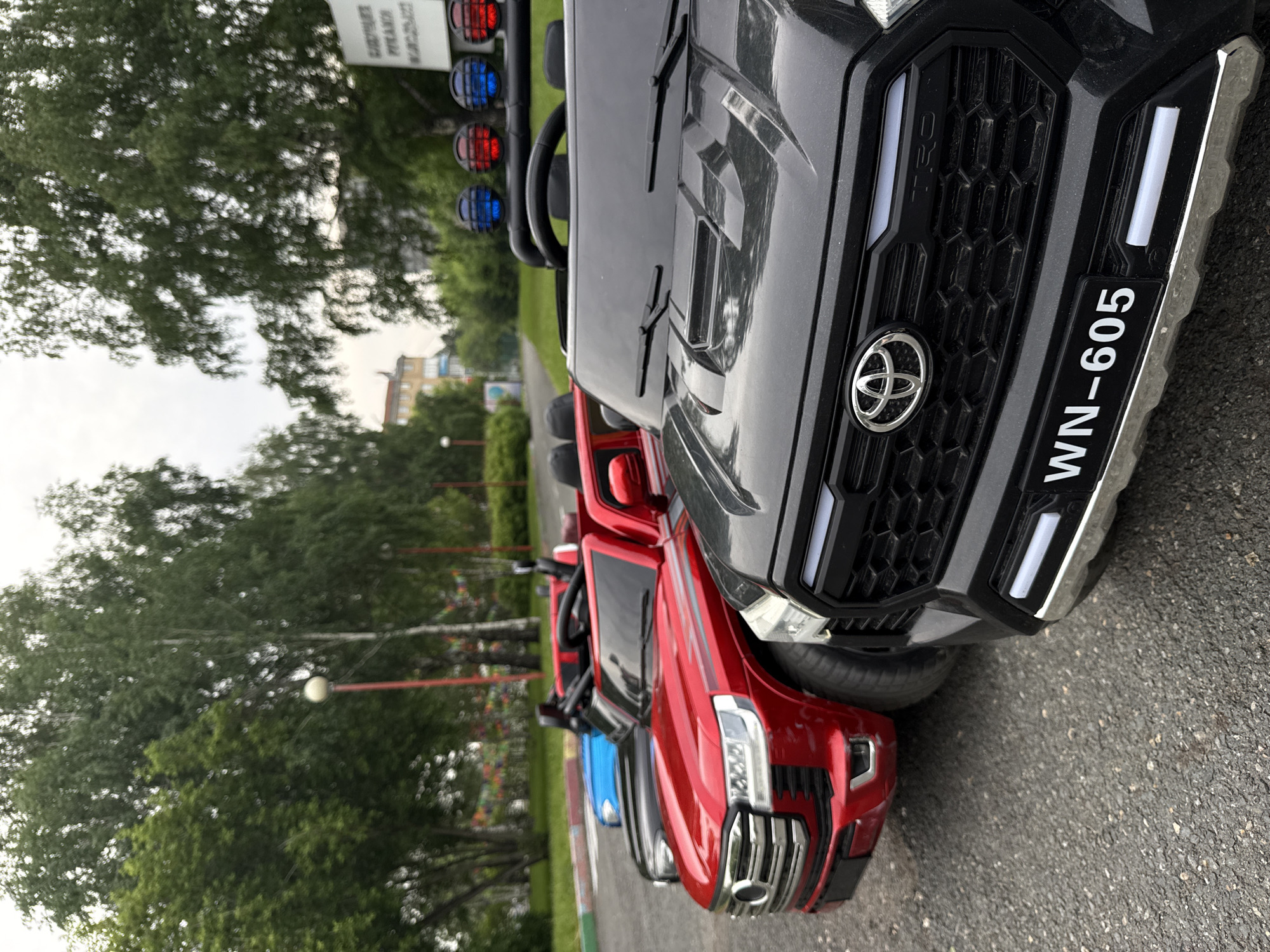 The image shows a row of electric ride-on toy cars parked on an asphalt surface outdoors. The cars are a red SUV, a black SUV with a license plate reading "WN-605", and a blue car, partially visible behind the red one. The black car has red and blue lights on its roof. In the background, there are trees, a street lamp, and a building. A sign with Russian text is visible on the right side of the image. The scene appears to be in a park or residential area. The location is identified as Krasnoyarsk, Russia.