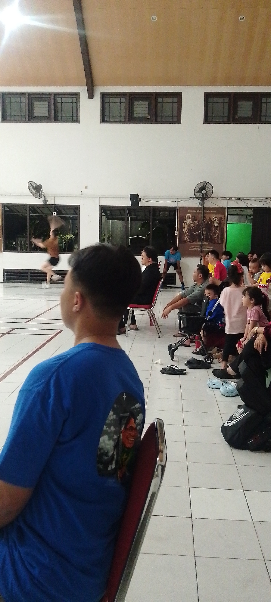 The image depicts an indoor event, possibly a performance or gathering, taking place in a tiled hall. In the background, a person is performing a dynamic movement, possibly dancing or participating in a martial art. Spectators are seated in chairs, facing the performance area. A prominent artwork depicting a religious scene hangs on the wall behind the seated audience. The hall has large windows, a ceiling fan, and fluorescent lighting. The foreground is occupied by a person in a blue t-shirt with a printed image on the back. The floor is covered with white square tiles, and various shoes are scattered around the seating area, suggesting a casual environment. The overall scene conveys a sense of community engagement and cultural activity. The location is identified as Kudus, Indonesia.