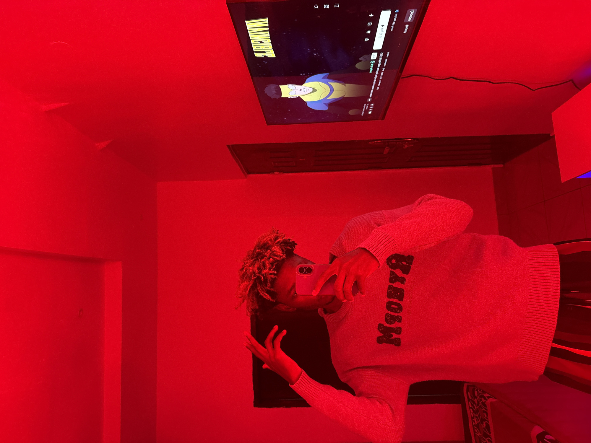 A person stands in a red-lit room, apparently taking a selfie with a cell phone. The person wears a gray sweater with black lettering. Overhead, a television displays an animated show. The setting is a room interior, likely in Igboefon, Nigeria.