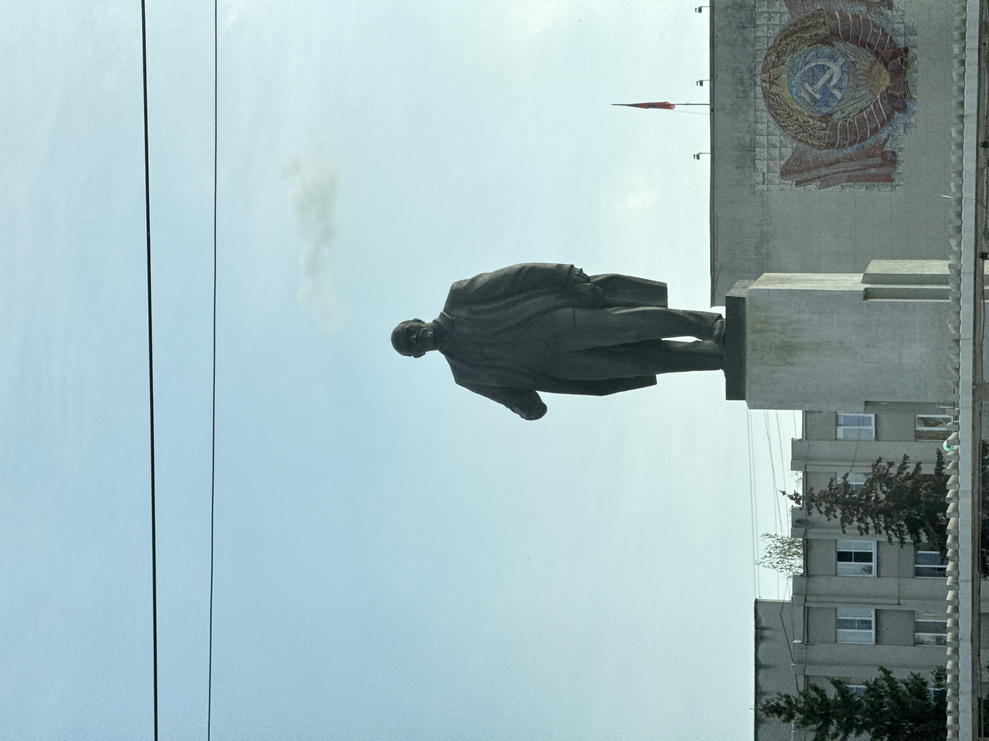 The image depicts a statue of Vladimir Lenin in Kstovo, Russia, standing in front of a building adorned with a Soviet-era emblem. The statue is facing left in the frame and elevated on a concrete platform. A red flag can be seen on top of the building with the emblem. Electrical wires are visible in the sky on the left of the image. An apartment building is adjacent to the statue. The sky is light blue.