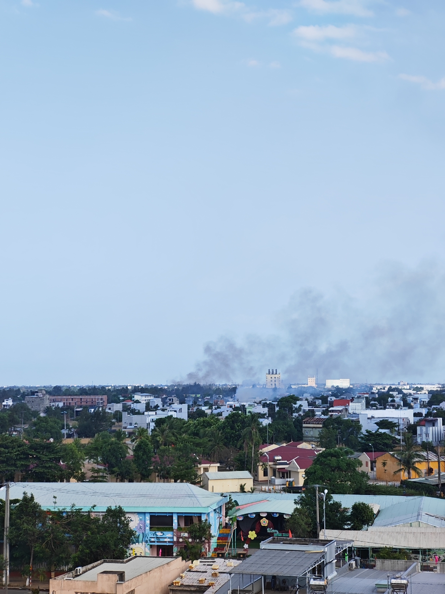 An aerial perspective captures a hazy urban landscape under a pale blue sky with sparse clouds. Smoke plumes rise from the mid-ground, obscuring a cluster of buildings, including what appear to be industrial structures with chimneys in the distance. The foreground is dominated by the roofs of various buildings, including what seems to be a school with colorful murals on its facade. Residential and commercial structures with diverse roof styles are interspersed with lush green trees throughout the city. The scene suggests a location in Đà Nẵng, Vietnam, given the architectural style and vegetation.
