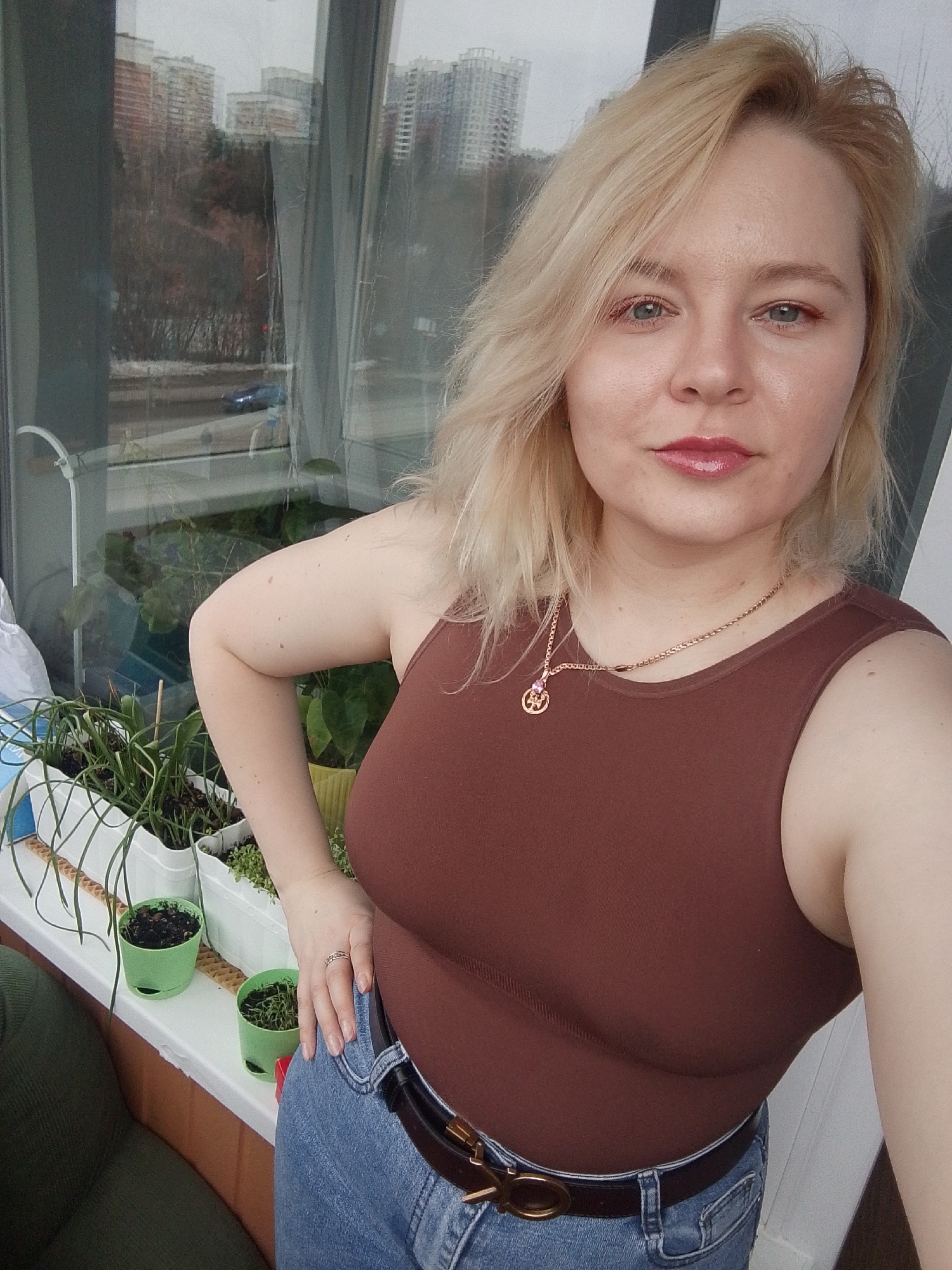 A woman poses for a selfie on a balcony in Izhevsk, Russia. She has blond hair, blue eyes, and is wearing a brown tank top, jeans, and a gold necklace. She has a ring on her finger. She has her hand on her hip. On the balcony railing are several potted plants. Through the window, a city skyline is visible, with a road and trees in the foreground. The weather appears overcast.