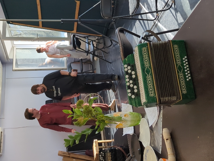 This image depicts an indoor scene in Moscow, Russia, resembling a music studio or rehearsal space, featuring three adult males and various musical instruments and objects.
In the foreground, on a dark table, a green Russian garmon (accordion) with white buttons and decorative patterns is prominently positioned. Adjacent to it are a white plastic recorder, several white ceramic plates, an orange cup, sheets of paper with text, and a black frying pan. A transparent glass vase containing green leafy branches stands near these items.
Three adult males are visible in the mid-ground. On the left, a man with brown hair and glasses, wearing a maroon long-sleeved shirt and a necklace, stands facing a bearded man. The bearded man, wearing a black t-shirt with white Cyrillic text "Мусорийский Кузнец" (Musoritsky Kuznets) and dark trousers, holds a small brown object in his right hand. Both appear to be engaged in conversation or standing in proximity. To the right, a third, younger male with short hair, dressed in a white tank top and light grey sweatpants, stands near a large, partially open window, with his left elbow on the windowsill and his right hand near his mouth, looking outwards.
The room has light-colored walls and a dark floor. Bright natural light streams through the window, illuminating bare tree branches outside and casting distinct shadows indoors. An air conditioning unit is mounted on the far wall. Additional items visible include a dark accordion case, a small wooden stool, stacked cardboard boxes, a black speaker, a partially obscured wheelchair, black folding chairs, and a large dark panel on a tripod stand.