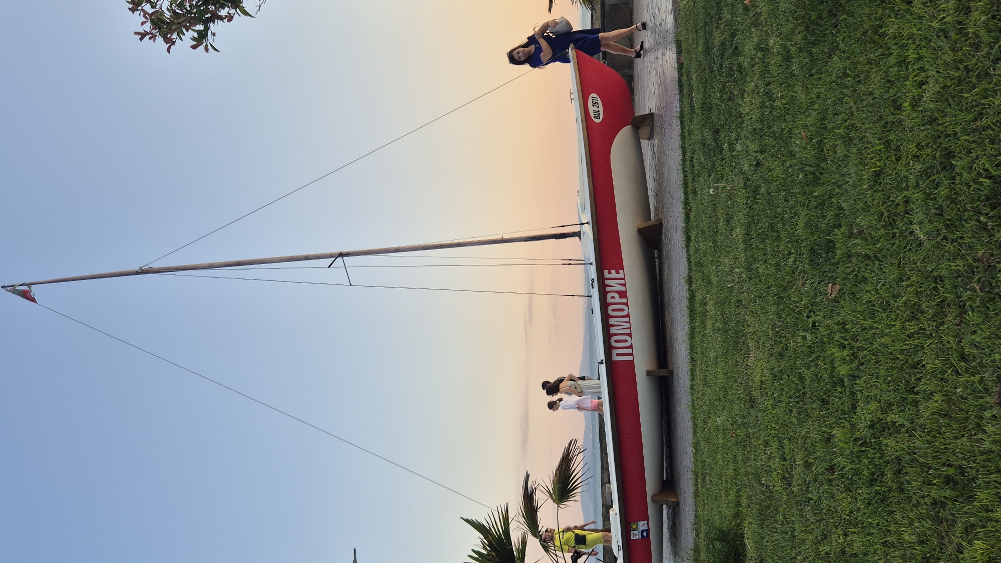 The image depicts a red and white sailboat on display near a waterfront in Pomorie, Bulgaria. The boat, with the word "ПОМОРИЕ" (Pomorie) emblazoned on its side, rests on wooden supports on a paved walkway. To the left of the boat, a person in a bright yellow outfit is visible. Several other individuals are standing near the boat, some looking out towards the sea. On the right, a woman in a blue dress is walking away from the boat, carrying a bag. Behind the boat, the calm sea meets the sky, with the horizon line faintly visible. The sky exhibits a soft gradient of colors, transitioning from blue at the top to a pale orange and pink near the horizon, suggesting it is either sunrise or sunset. A tall flagpole with a small flag stands behind the boat, with wires extending from it. The foreground is covered with lush green grass. The scene appears to be a public space, possibly a promenade or park in the coastal town of Pomorie.