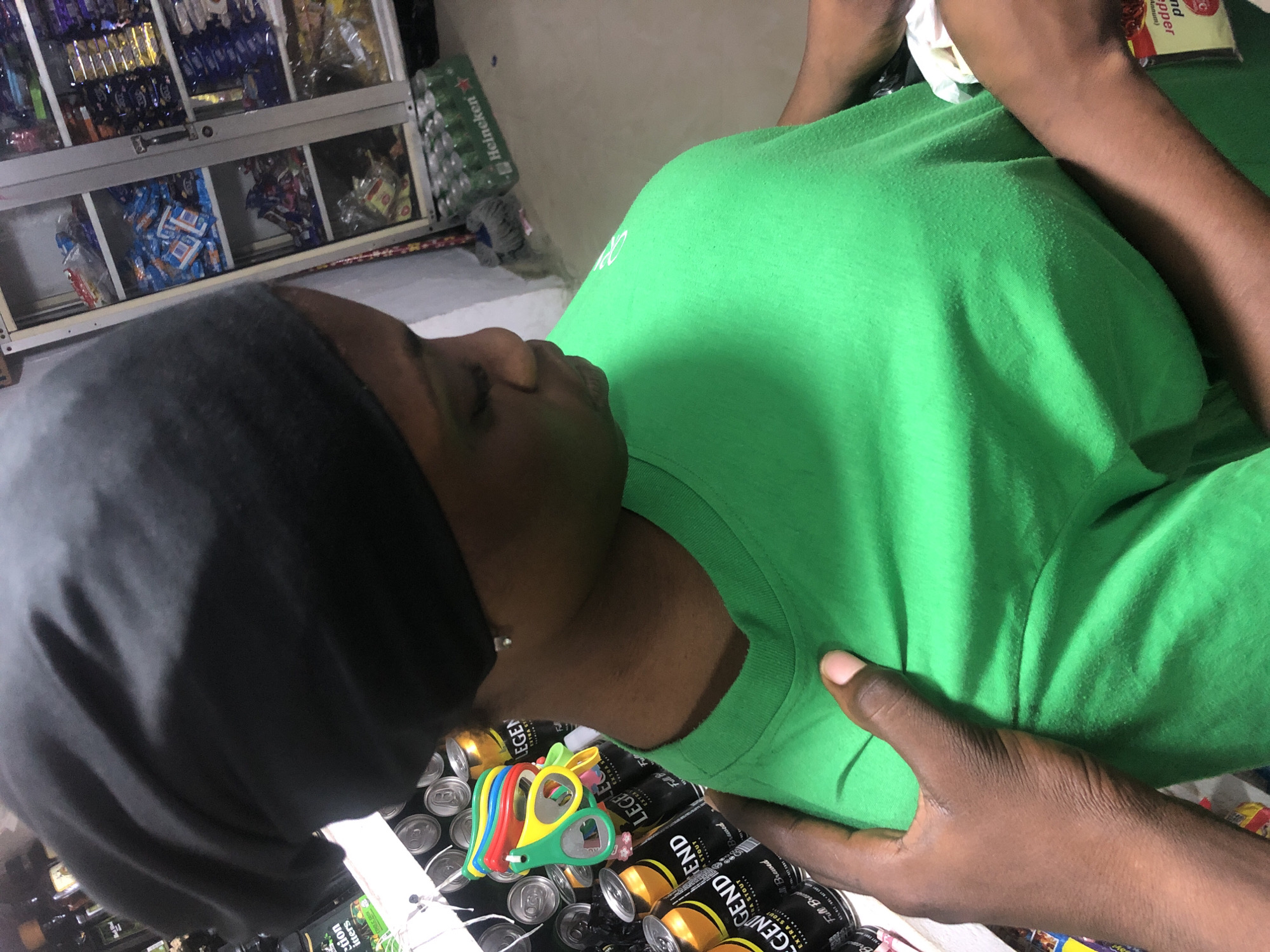 A medium shot captured from an elevated angle shows a person in a store in Ado, Nigeria. The person, with dark skin and wearing a black headscarf and a vibrant green t-shirt, is positioned centrally and facing away from the camera, looking down. Their right hand is visible, placed on their own chest.
The background reveals shelves stocked with various goods, including what appear to be packaged snacks and drinks. Bottles of "Legend Extra Stout" are prominently displayed on a shelf behind the person. A case of Heineken beer is visible on the floor in the lower right portion of the frame. The store appears to be a small retail establishment, typical of those found in Nigerian cities. The lighting is somewhat dim, suggesting indoor illumination.