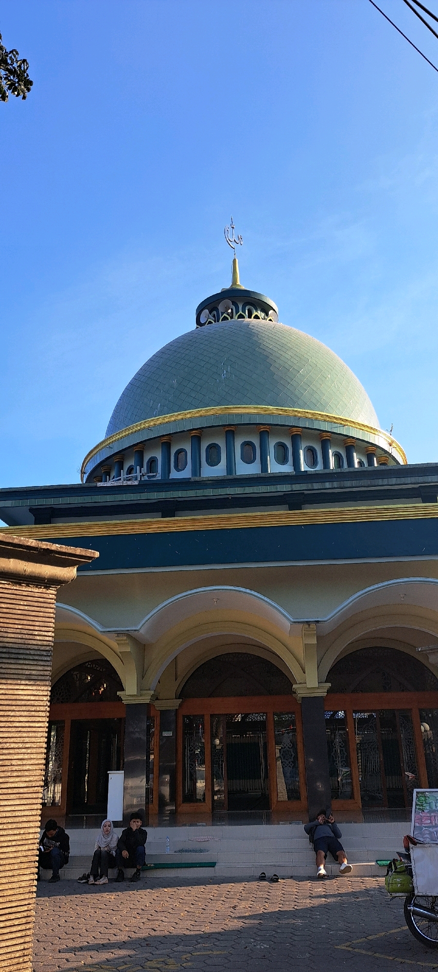 A low-angle, full-shot photograph captures a mosque in Gambiran, Indonesia, under a clear blue sky. The mosque features a large, greenish-blue dome with gold trim, topped by a golden spire with an Arabic inscription. A band of decorative circular windows encircles the base of the dome, supported by pillars. Below the dome, the mosque's facade is a lighter color, with arched entrances and dark wooden doors. To the left, a textured, brick-like wall partially obscures the view. On the steps leading to the mosque, several individuals are seated or resting. In the foreground, a paved area with a repeating diamond pattern is visible. A small cart with a green bag is parked on the right side of the image.