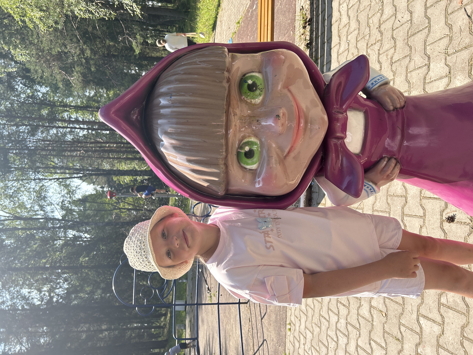 In Krasnovishersk, Russia, a young girl wearing a straw hat, a pink t-shirt, and white shorts poses next to a large statue of the cartoon character Masha. The statue is vibrantly colored in shades of pink and purple. The girl is looking towards the camera with a slight smile. Behind them, a park scene unfolds with trees, a playground structure, and a paved walkway. Sunlight filters through the trees, indicating it is daytime. Another person can be seen in the background walking away from the camera. The ground is paved with interlocking concrete bricks.