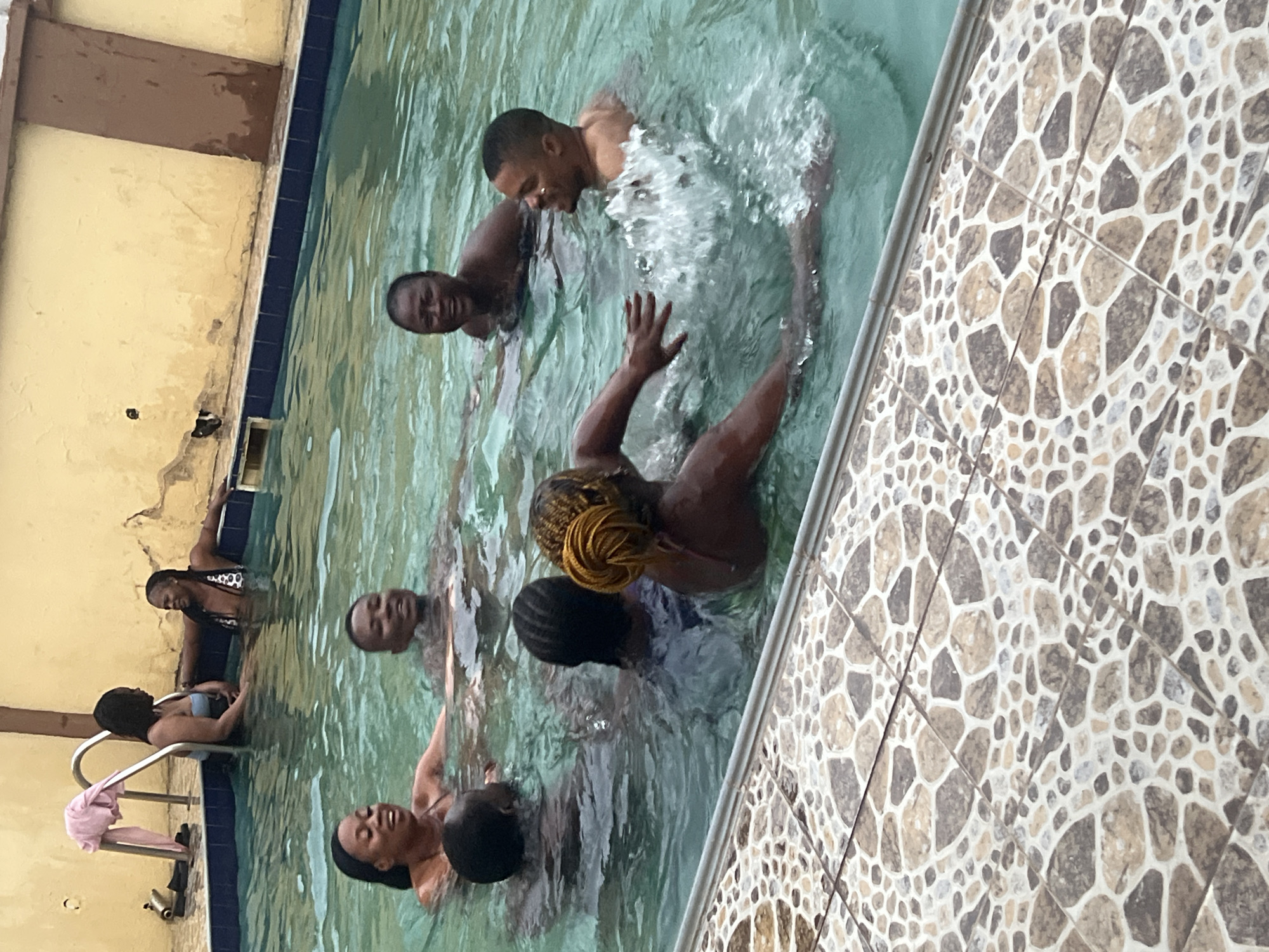Several people are in a swimming pool. The pool is surrounded by tiled flooring and a wall with peeling paint. Some individuals are actively swimming while others are closer to the pool's edge. The image appears to have been taken at an angle. The exact location within Abuja, Nigeria, is indiscernible from the image alone.