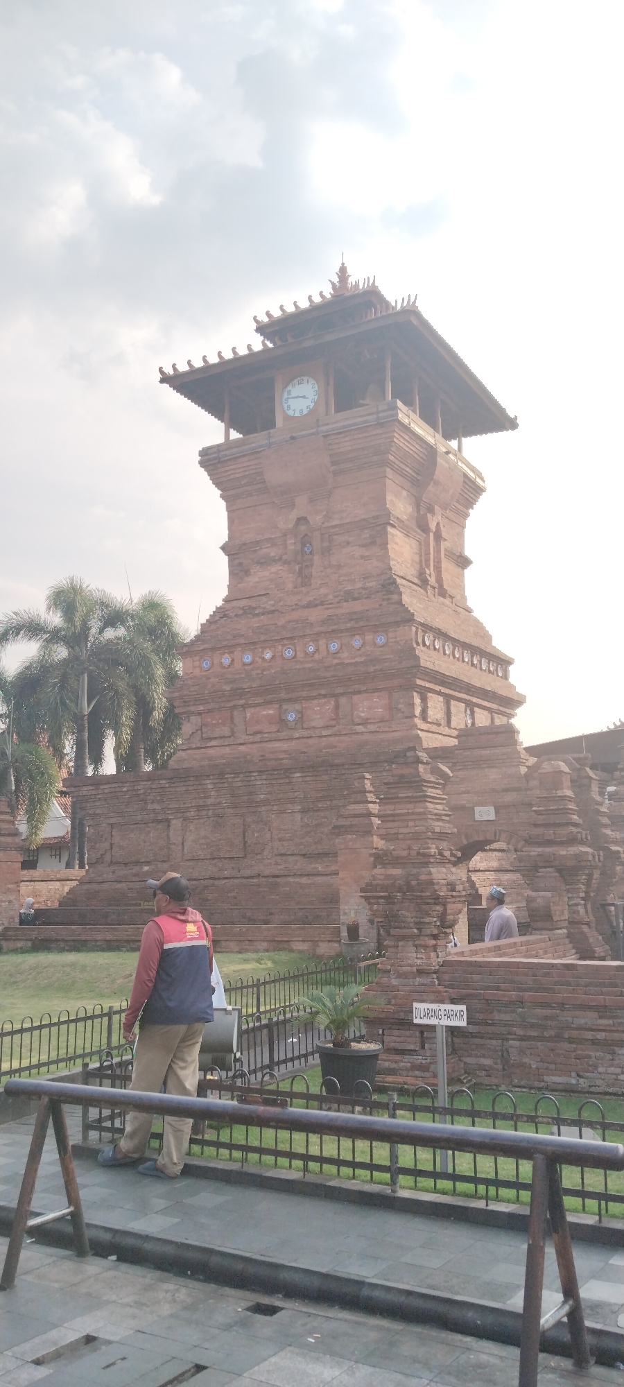 The image shows the Menara Kudus Mosque in Kauman, Indonesia. The mosque is a historic landmark known for its unique architecture, which blends Islamic and Hindu influences. The tower, made of red brick, has a square base and a tiered roof. A clock is visible on one side of the tower, and decorative elements are present on the brickwork. In the foreground, a man wearing a vest and a cap is standing near a metal fence. Palm trees are visible to the left of the tower. The sky is cloudy.