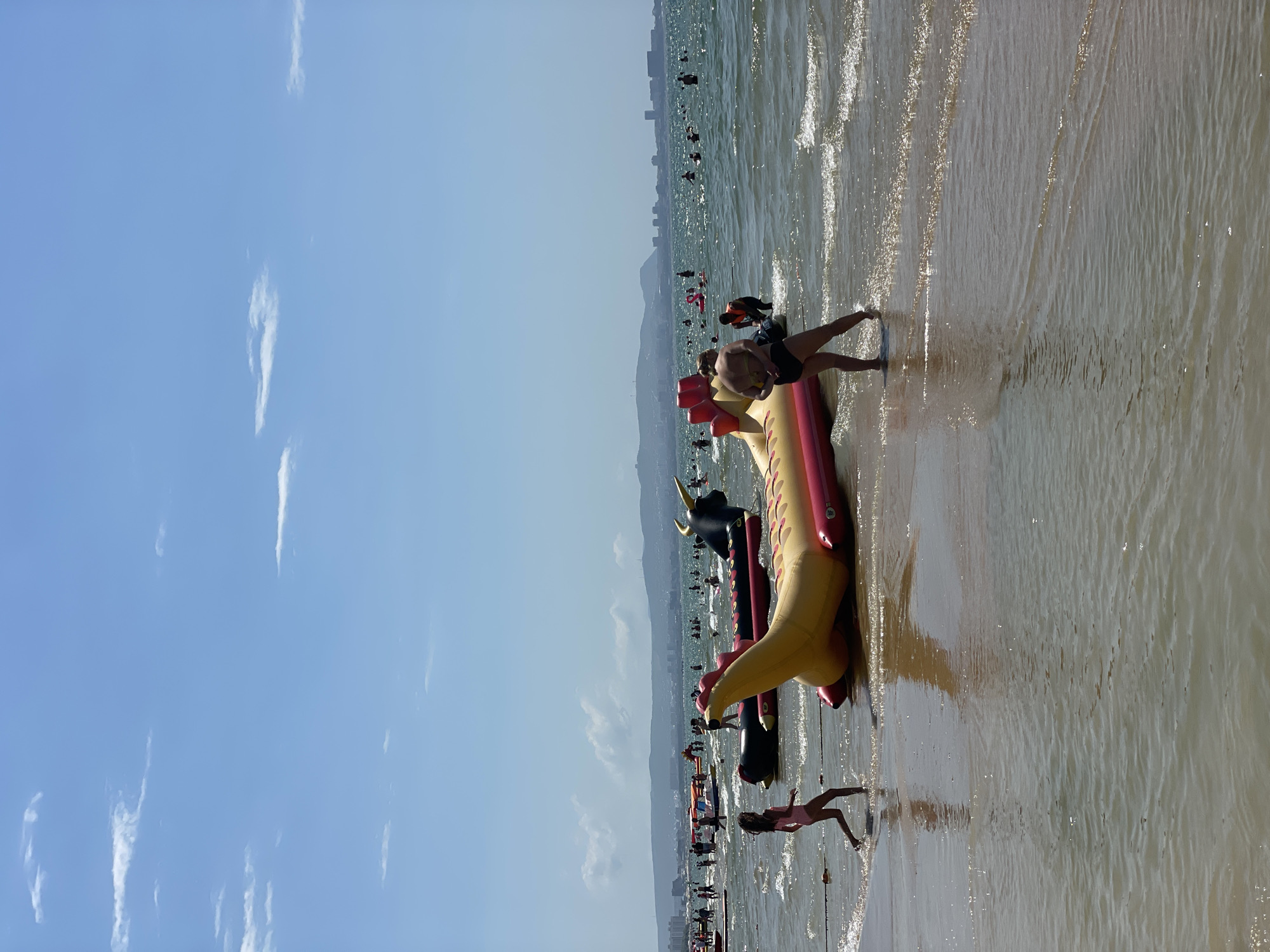 The image depicts a crowded beach in Anapa, Russia on a sunny day. The foreground is dominated by shallow, rippling water with reflections of the sky. In the middle ground, several inflatable water toys are present. A prominent yellow and red inflatable serpent-like toy is positioned in the center, with two individuals standing near it. To the left, a young girl in a pink swimsuit is walking through the water. Further out in the sea, numerous people are visible swimming and enjoying various water activities. In the background, a cityscape with buildings and hills lines the horizon under a partly cloudy blue sky.
