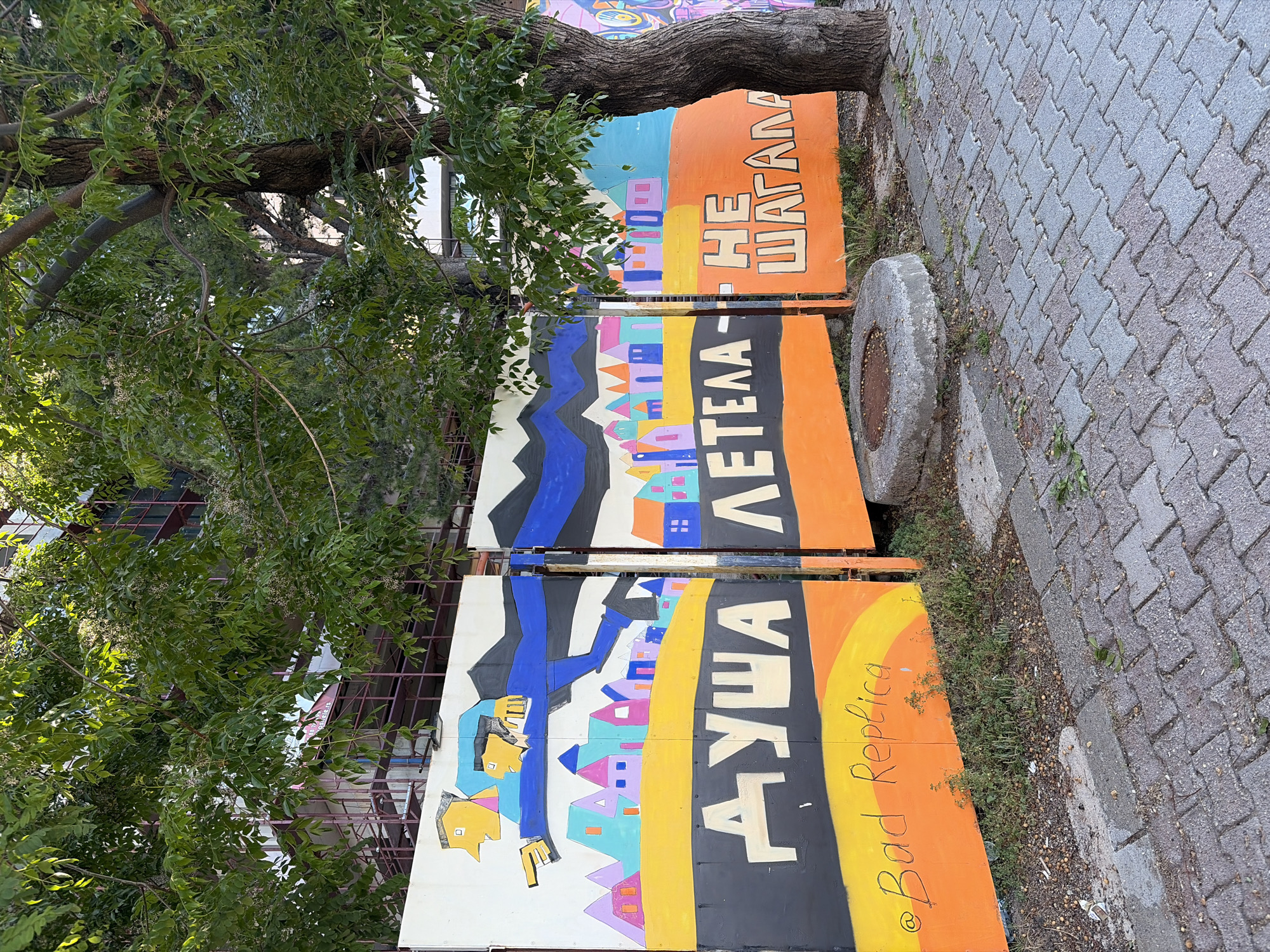 The image depicts a street scene in Rafailovići, Montenegro. Three brightly colored, vertically oriented painted panels are positioned near a tree and a paved walkway. The panels feature stylized, illustrative designs with text in a Cyrillic alphabet. The bottom panel depicts a yellow face in profile above colorful buildings. The middle panel shows colorful buildings under dark mountains, and the top panel is similar. "@Bad Replica" is written on the bottom panel. The background includes foliage from the tree and glimpses of buildings. The walkway is made of light grey paving stones. A circular concrete object is adjacent to the top panel.