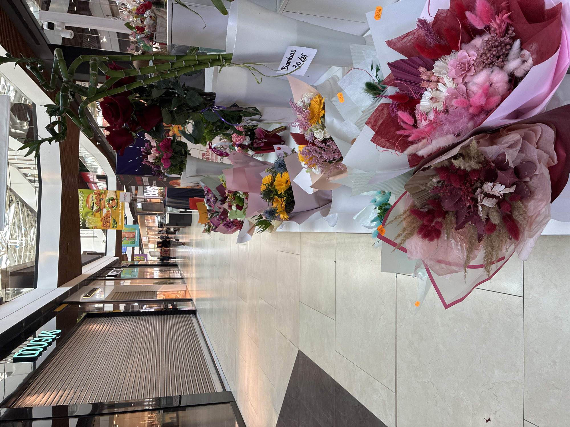 The image depicts a shopping mall interior, likely in Brasov, Romania, given the presence of Romanian currency prices on some items. On the left, a row of closed shopfronts is visible, one with a prominent "DOUGLAS" sign. The floor is tiled in a light beige color. In the foreground and mid-ground, a flower stall displays a variety of bouquets and plants. Several white vases hold arrangements of flowers and bamboo stalks. One sign reads "Bambus 30 lei". The bouquets vary in color and composition, featuring dried flowers, roses, and other decorative elements. The overall lighting suggests an indoor environment with overhead illumination. In the background, shoppers can be seen walking down the mall. A large banner featuring a burger advertisement is visible further down the corridor.