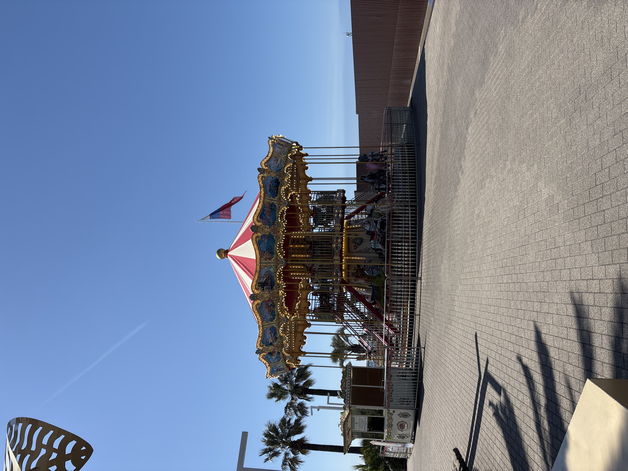 Here's a detailed description of the image:
The image shows a brightly lit, two-tiered carousel against a clear blue sky. The carousel is decorated with gold trim and has a red and white striped roof topped with a flag that appears to be the Czech flag. The ride is enclosed by a metal fence. Behind the carousel are palm trees and part of a building with a textured wall. The ground is paved with gray bricks. Shadows from the palm trees and another object (likely part of a structure) are visible on the pavement.
Based on the style of carousel and the presence of palm trees, it is highly plausible the image was taken in Sochi, Russia, due to its coastal location and the presence of common Russian symbols.