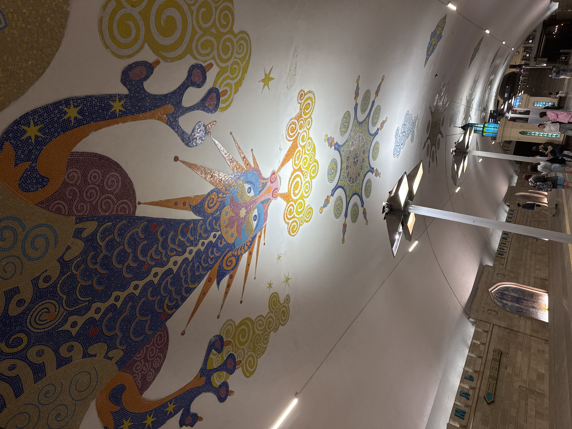 The image shows a mosaic mural on the ceiling of a subway station. The main subject is a colorful dragon-like creature made of small mosaic tiles, with a swirling pattern around it. The background is white. There are lights and a ceiling beam that intrude into the scene. In the background, there are people and details of the station interior. The location is Unknown, Unknown.