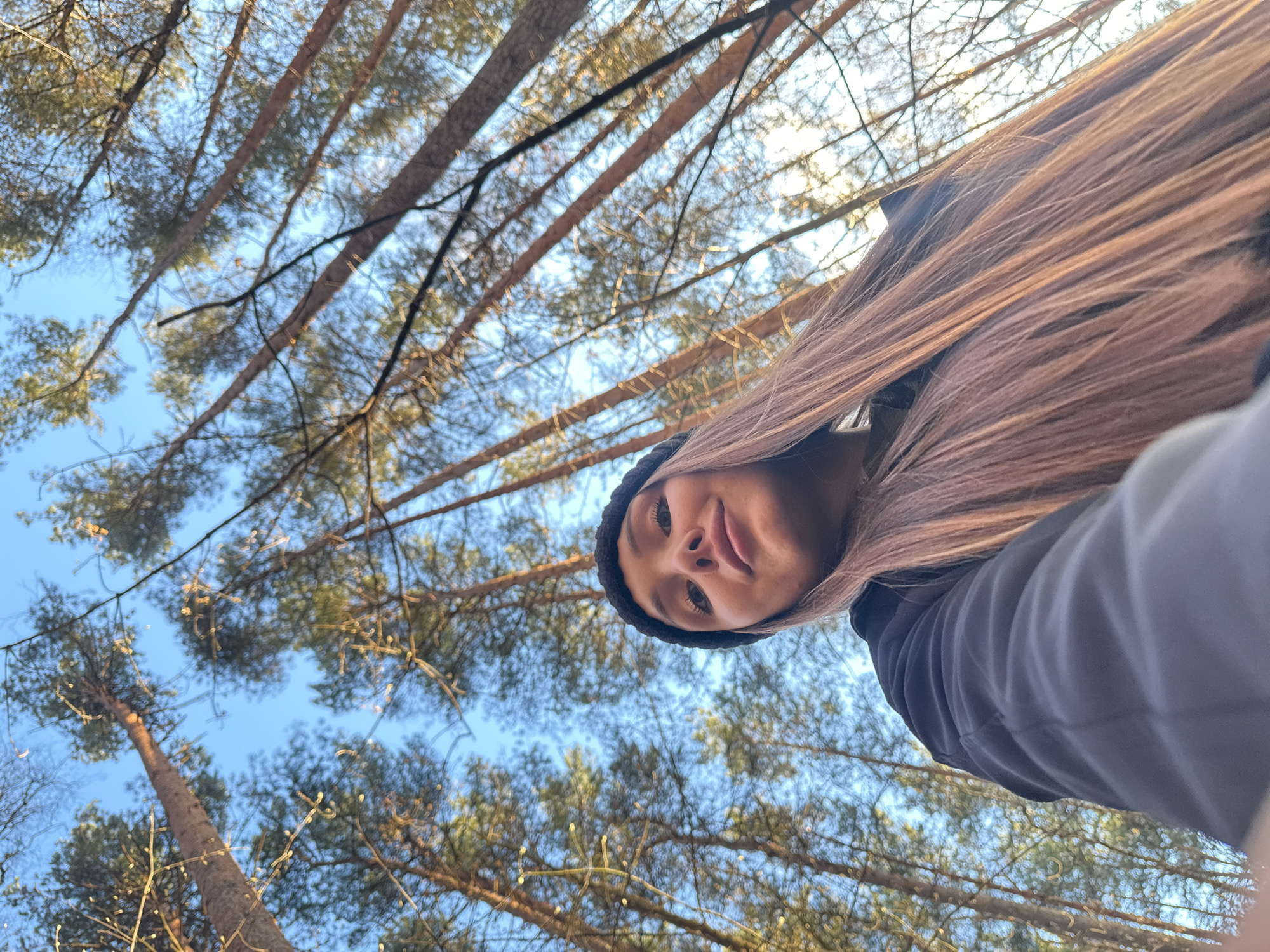 A low-angle selfie captures a young woman looking down into the camera with a forest canopy and clear blue sky as the backdrop. The woman is positioned in the upper right quadrant of the frame, with her light brown hair flowing towards the right. She wears a dark beanie and a dark jacket, and has a neutral to slightly serene expression, making direct eye contact with the viewer.
The majority of the image features the upward view into a dense pine forest, with numerous tall, straight tree trunks and branches extending across the clear, bright blue sky. Sunlight filters through the sparse needles, creating bright spots and a pleasant, airy atmosphere. Some faint, circular bokeh effects are visible among the branches, possibly from decorative lights or sunlight reflecting off water droplets. The overall impression is that of a bright, sunny day outdoors. The setting suggests a natural environment, likely a park or a wooded area in Chaikovsky, Russia. There is no visible text in the image.