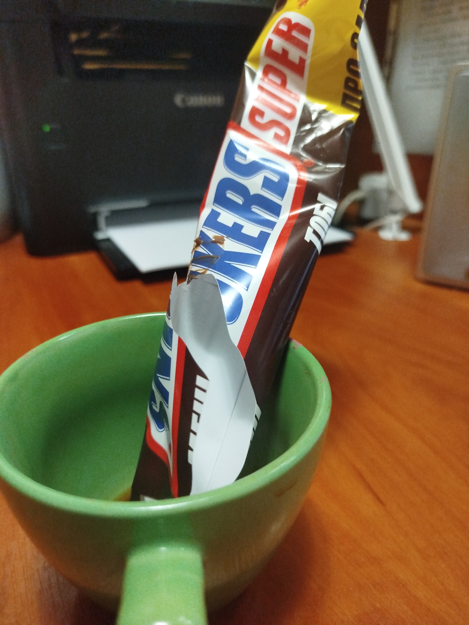 A partially unwrapped Snickers candy bar is placed inside a green mug. The candy bar, with its distinctive yellow, red, and blue packaging, is positioned vertically, with its wrapper torn open to reveal the chocolate inside. The mug itself is a vibrant green, with a handle visible. The background is blurred, but appears to be an indoor setting with a wooden desk or table. A black printer with a green indicator light is partially visible to the left, and other office equipment is suggested in the background. The overall scene is a close-up shot, focusing on the Snickers bar and mug. The image does not contain any information to determine its location in the city of Pavlohrad, Ukraine.