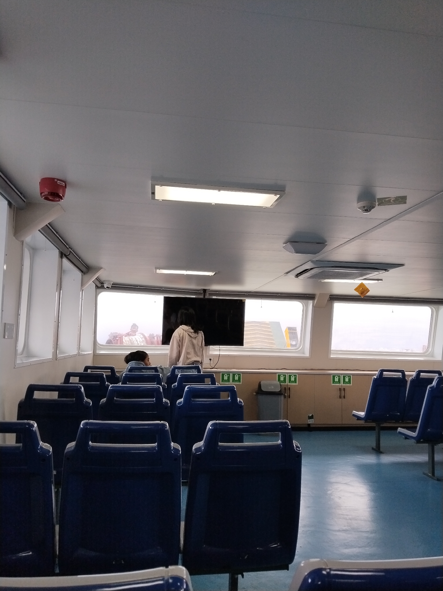 The image depicts the interior of a ferry cabin, likely in or near Melaya, Indonesia. The cabin is outfitted with rows of blue plastic seats. Large windows line the walls, offering a view of the exterior, which appears to be a hazy or foggy environment with structures visible in the distance. Several individuals are present in the cabin. In the foreground, the backs of multiple empty seats are visible. Further into the cabin, a person in a light-colored hooded sweatshirt is standing and looking towards a television screen mounted on the wall. Another person is seated, facing away from the camera. The ceiling features fluorescent lighting, a smoke detector, and what appears to be an air conditioning unit. Safety signage, including life preserver icons, is visible on the walls. A trash receptacle is also present. The overall environment suggests a functional passenger transport vessel.