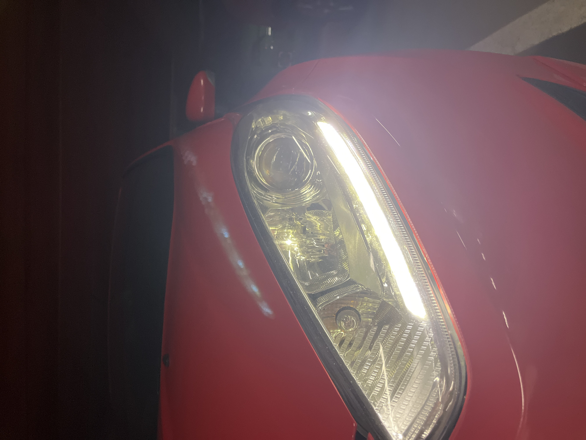 The image shows the front right headlight of a red car, likely a Ford Fiesta, at night. The headlight is illuminated, emitting a bright white light. The car's red paintwork is visible, reflecting some of the light. The background is dark and out of focus, suggesting an outdoor setting, possibly a street or parking lot. There is no indication of the specific location in Vantaa, Finland.