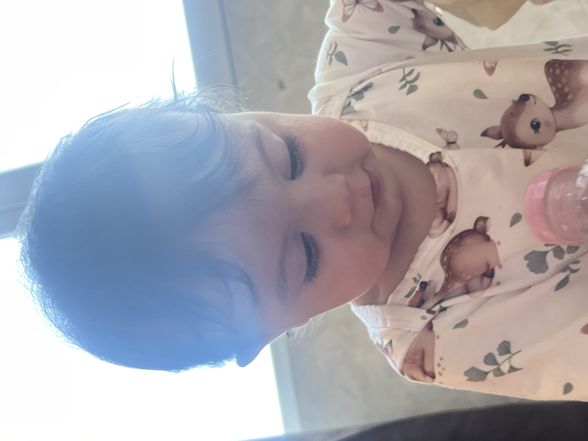 A close-up, slightly tilted, indoor shot shows a baby wearing a white outfit with a pattern of deer and leaves. The baby is lying down, with eyes closed and a slight smile. Part of a pink baby bottle is visible on the right. A bright light source is shining on the left side of the baby's head. The location is not discernible from the image.