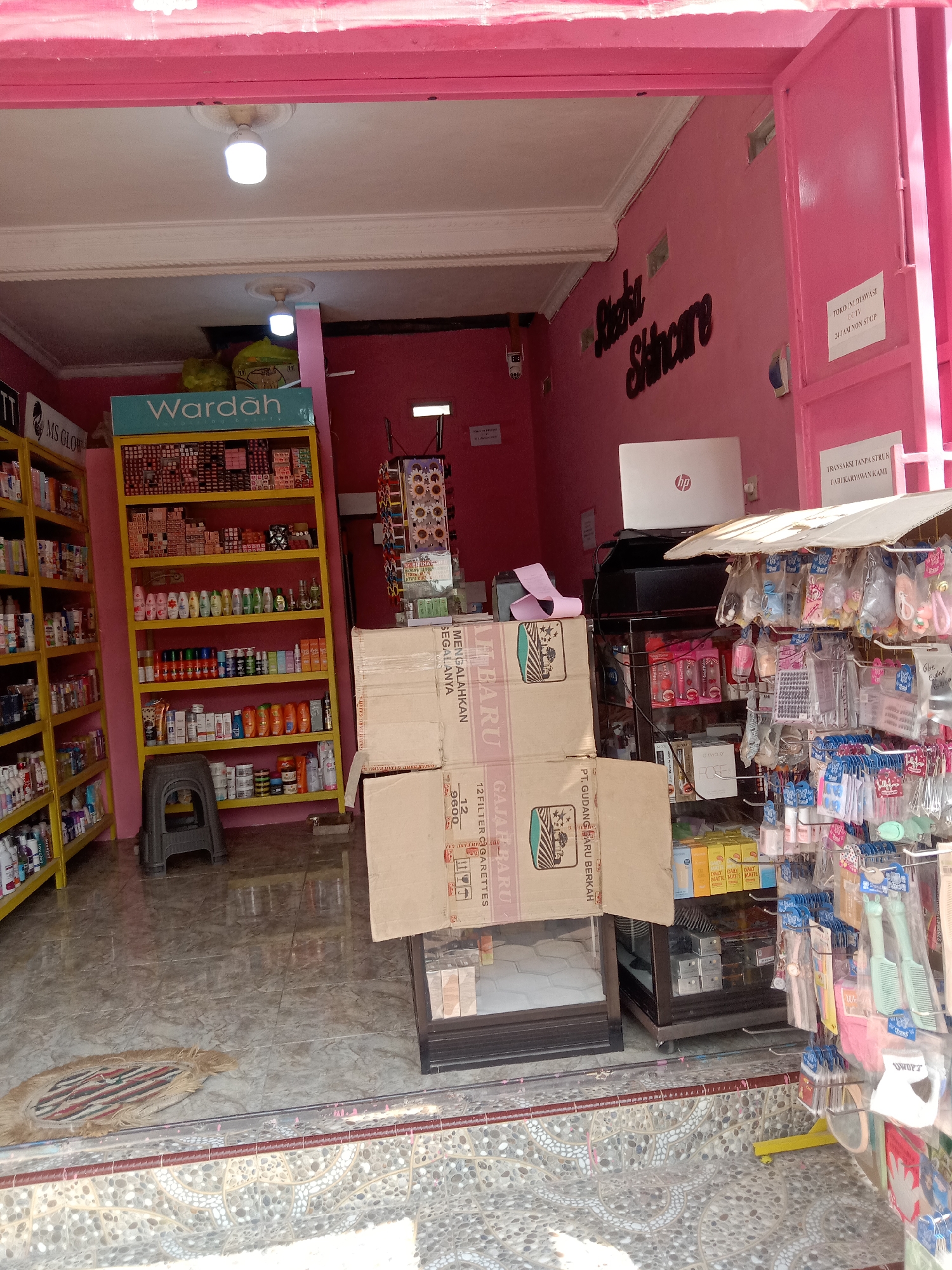 The image depicts the exterior of a small cosmetics store, likely a family-run business, located on a street in Sugihwaras, Indonesia. The store is painted a vibrant pink and features an open storefront displaying a variety of beauty products. A prominent "Wardah" branded shelf is visible, stocked with numerous skincare and makeup items. To the left, another shelving unit displays more products, and to the right, a rack hangs with various personal care items such as hairbrushes and cosmetic accessories. Cardboard boxes are stacked near the entrance, some with visible text in Indonesian. The overall impression is of a bustling, local business catering to the community's beauty needs. The pavement in front of the store is decorated with intricate tile work. No people are visible in the image.