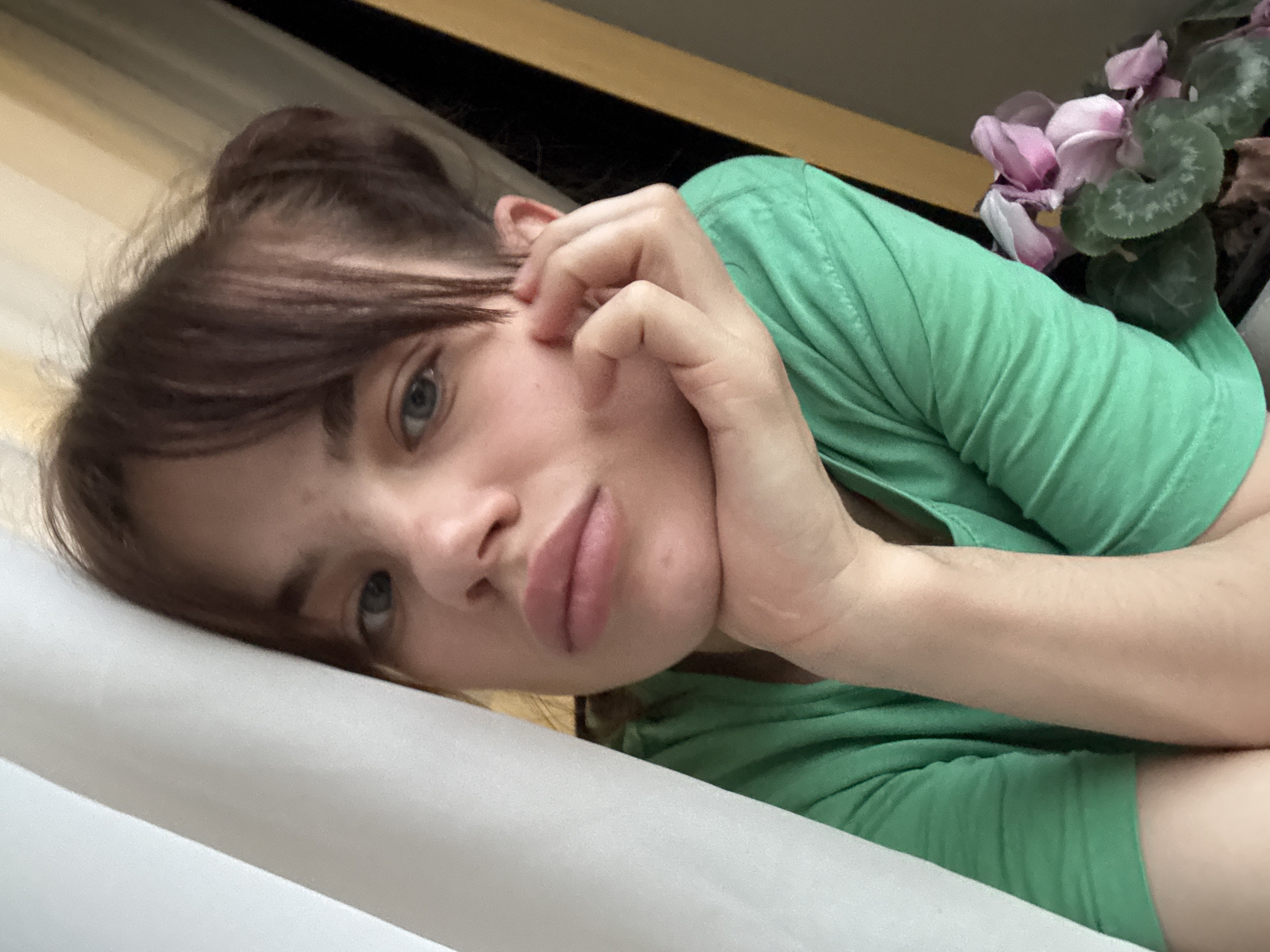 A woman lies on a white surface, possibly a bed or a couch. She wears a green t-shirt. Her brown hair is styled in a loose bun with bangs falling across her forehead. She rests her cheek on her hand, fingers gently touching her face. To the upper right of the frame, a potted plant with pink cyclamen flowers sits on a surface. The location of Rostov-on-Don, Russia, is not discernible from the image.
