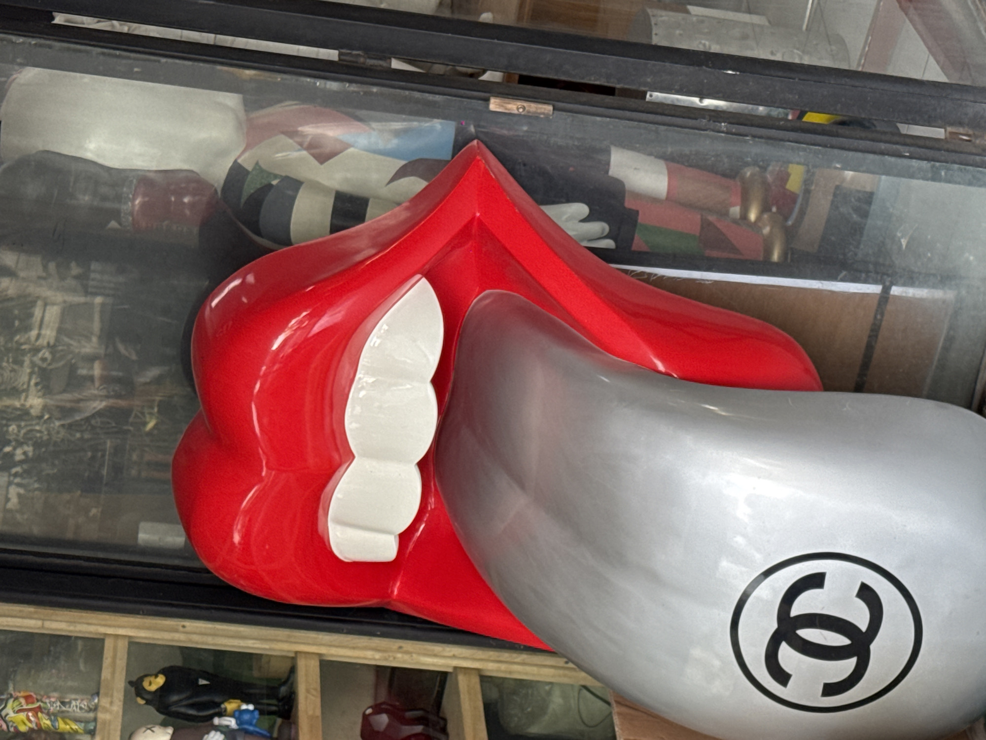 An outdoor, eye-level, medium shot captures a vibrant red sculpture of lips with a silver tongue, emblazoned with a black Chanel logo. The sculpture is positioned in front of a glass shop window in North Kuta, Indonesia. Behind the lips, reflections in the glass reveal a display of mannequins with colorful patterned clothing and gold shoes. To the left of the sculpture, shelves are visible, holding various figurines, including a black monkey-like figure and a white cartoonish figure. The lighting appears to be natural daylight.