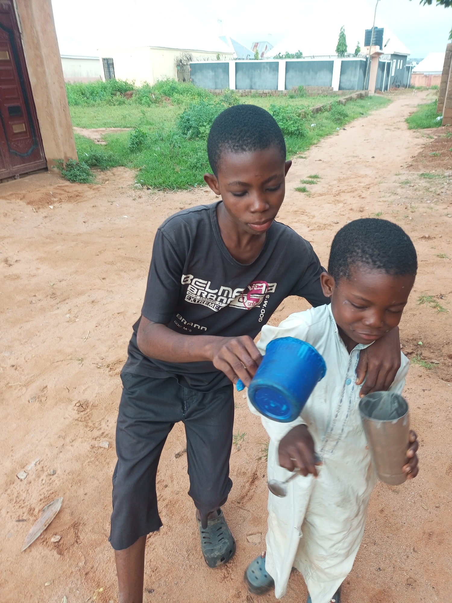 Two young boys are standing in a dirt area outside. The boy on the left, wearing a black t-shirt and black pants, is holding a blue cup with a small ladle in it. He is extending the ladle towards the boy on the right. The boy on the right is wearing a light-colored traditional garment and is holding a metal cup in his right hand. Both boys are looking down at the objects they are holding. The background shows a dirt path, some greenery, and buildings in the distance, suggesting an outdoor setting in a residential or semi-rural area.
