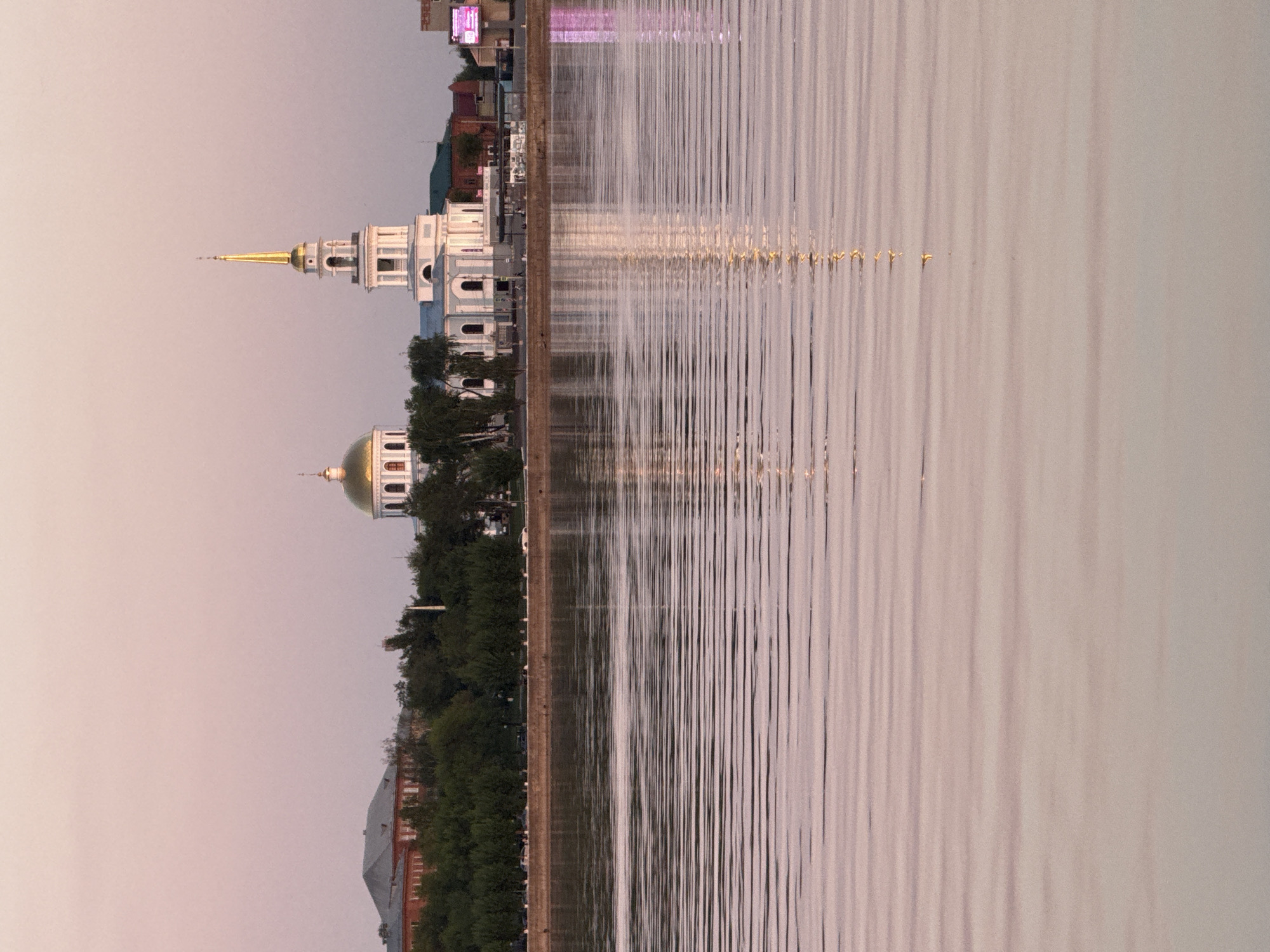 The image shows a waterfront scene in Votkinsk, Russia. On the left side of the image are structures including a domed religious building with a golden dome, another building with a golden spire, and trees lining the waterfront. The right side of the image shows the calm water reflecting the structures and trees. The sky is a pale color. The image is rotated 90 degrees counterclockwise.