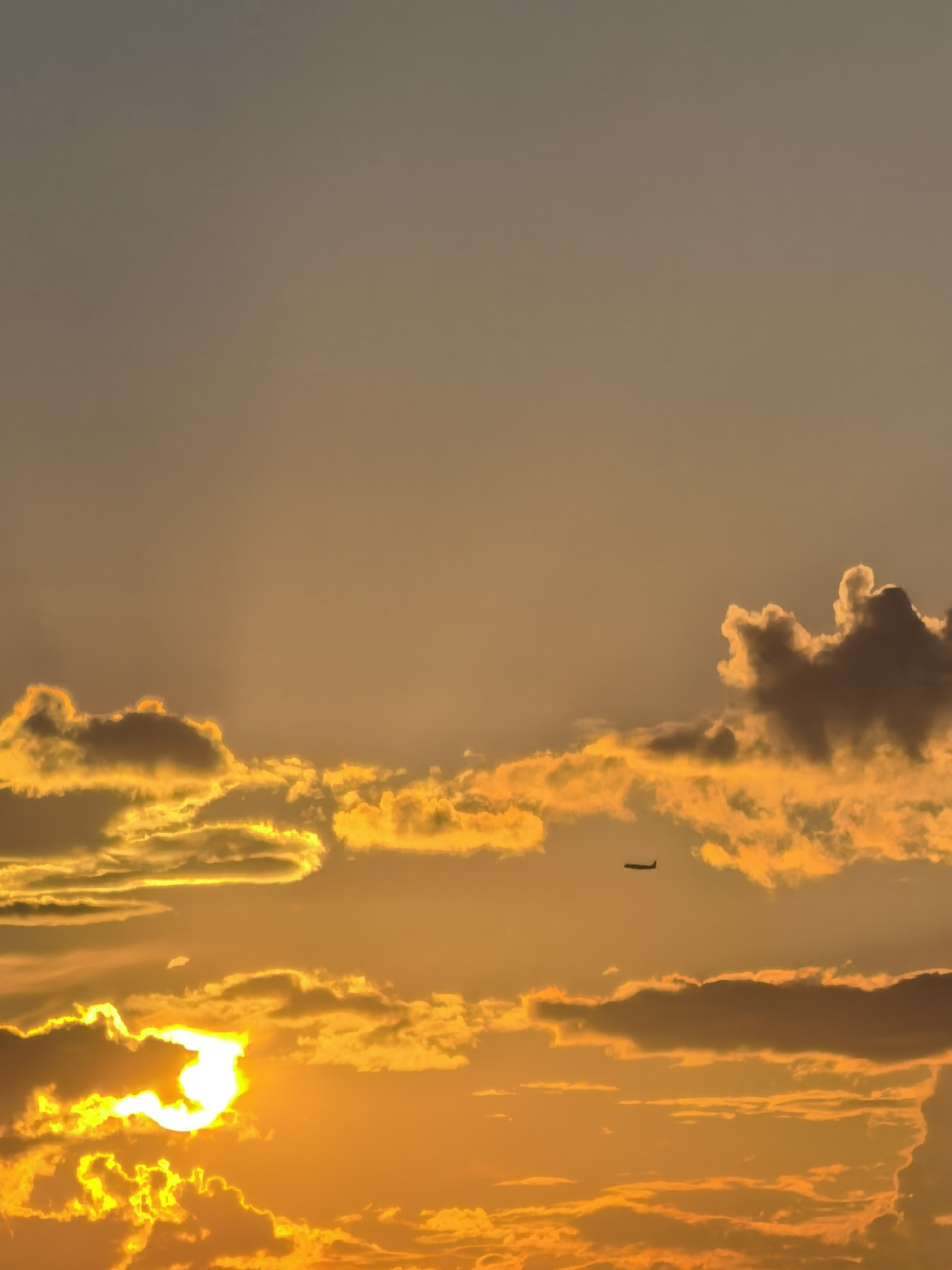 An aerial shot captures a sunset with vibrant orange and yellow hues. A lone airplane is silhouetted against the sky, cruising amongst the clouds. The sun, a brilliant white orb, peeks through a gap in the clouds, casting a warm glow on the scene. The image, taken from an unknown location in the city of Unknown, Unknown, depicts a serene and picturesque end to the day.