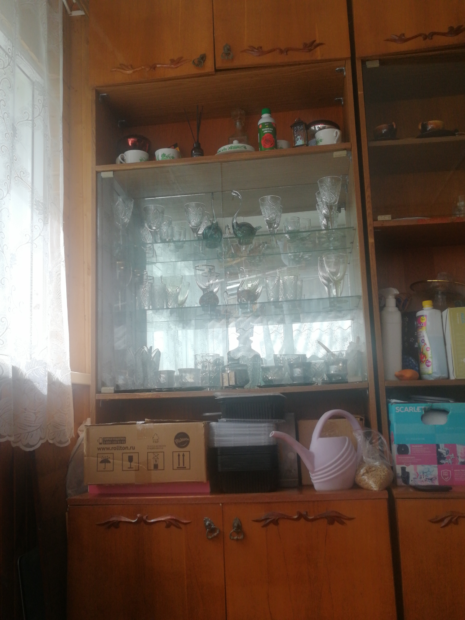 A close-up, eye-level shot captures a cluttered wooden cabinet in what appears to be a domestic setting. The cabinet is filled with an assortment of glassware, bottles, and boxes. On the top shelf, a green bottle with a red rose label is visible, alongside other small containers and a ceramic cup. The middle section features numerous clear glass items, including wine glasses, tumblers, and decorative pieces, arranged on glass shelves. Below, a cardboard box with Russian text and shipping symbols sits on a pink surface. To the right of this box, a light pink watering can and a plastic bag filled with what appears to be grain are placed on a shelf. Further to the right, a blue box with "SCARLETT" branding is visible. The overall impression is one of storage and organization of various household items within a wooden cabinet. The scene is likely located in Dubrovka, Russia.
