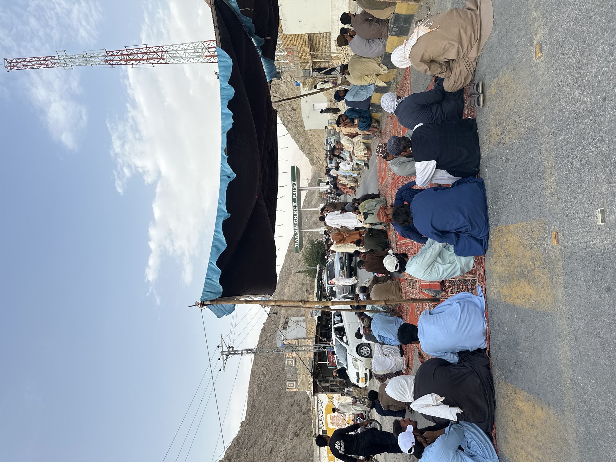 This image depicts a gathering of approximately 60-70 men sitting on carpets on an asphalt road, likely in the Tehsil of Quetta City, Pakistan, given the presence of a "HANNA CHECK POST" sign. The men, dressed in traditional Pakistani attire, are seated in rows, facing various directions, some in conversation, others looking towards the road. They appear to be participating in a peaceful protest or demonstration, as indicated by the presence of a large canopy providing shade, suggesting an extended gathering.
The environment is semi-arid, with barren hills visible in the background. Power lines and a prominent telecommunications tower are also present, indicating an established infrastructure. Several vehicles, including cars and motorcycles, are parked or driving on the road, suggesting this is a main thoroughfare. The overall atmosphere appears calm, with the focus on the seated assembly of men. The presence of the check post sign, a communication tower, and the general landscape point towards a border or a significant checkpoint area within the region.