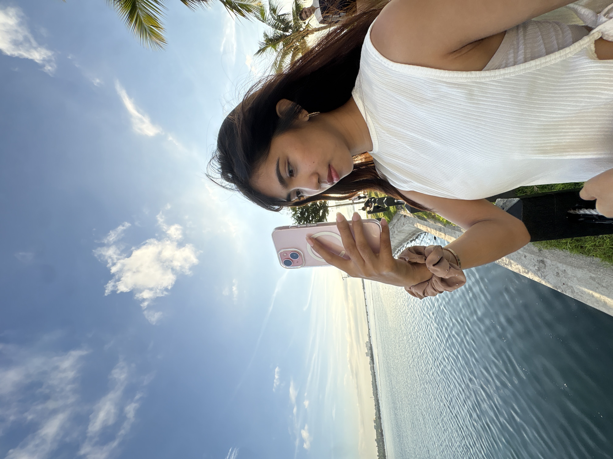 In Davao City, Philippines, a young woman takes a selfie by the coast. She is holding a pink iPhone in her right hand, which is adorned with a scrunchie on her wrist. The woman is wearing a white sleeveless top. Behind her, the calm blue water stretches towards a distant city skyline, bridged by a structure. The sky above is a clear blue, dotted with scattered white clouds. Palm trees line the background, suggesting a tropical setting. The scene appears to be captured during the day, with the sun casting a warm light.