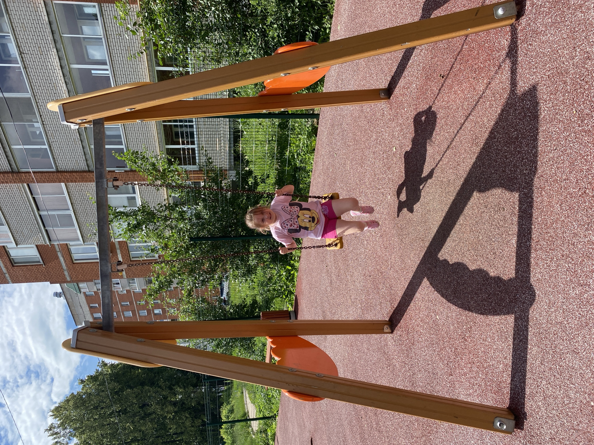 A young girl with blonde hair is swinging on a playground swing. She is wearing a pink t-shirt with a cartoon mouse graphic and pink shorts. The swing set is made of wood with orange accents. The ground beneath the swing is a red, rubberized surface, common in playgrounds. In the background, there is a tall brick building with many windows, green trees, and a chain-link fence. The sun is casting a shadow of the swing set and a person on the ground, indicating it is daytime with clear weather. The girl appears to be enjoying herself, with a slight smile on her face. The scene takes place outdoors at a playground, likely in a residential area given the presence of the apartment building.