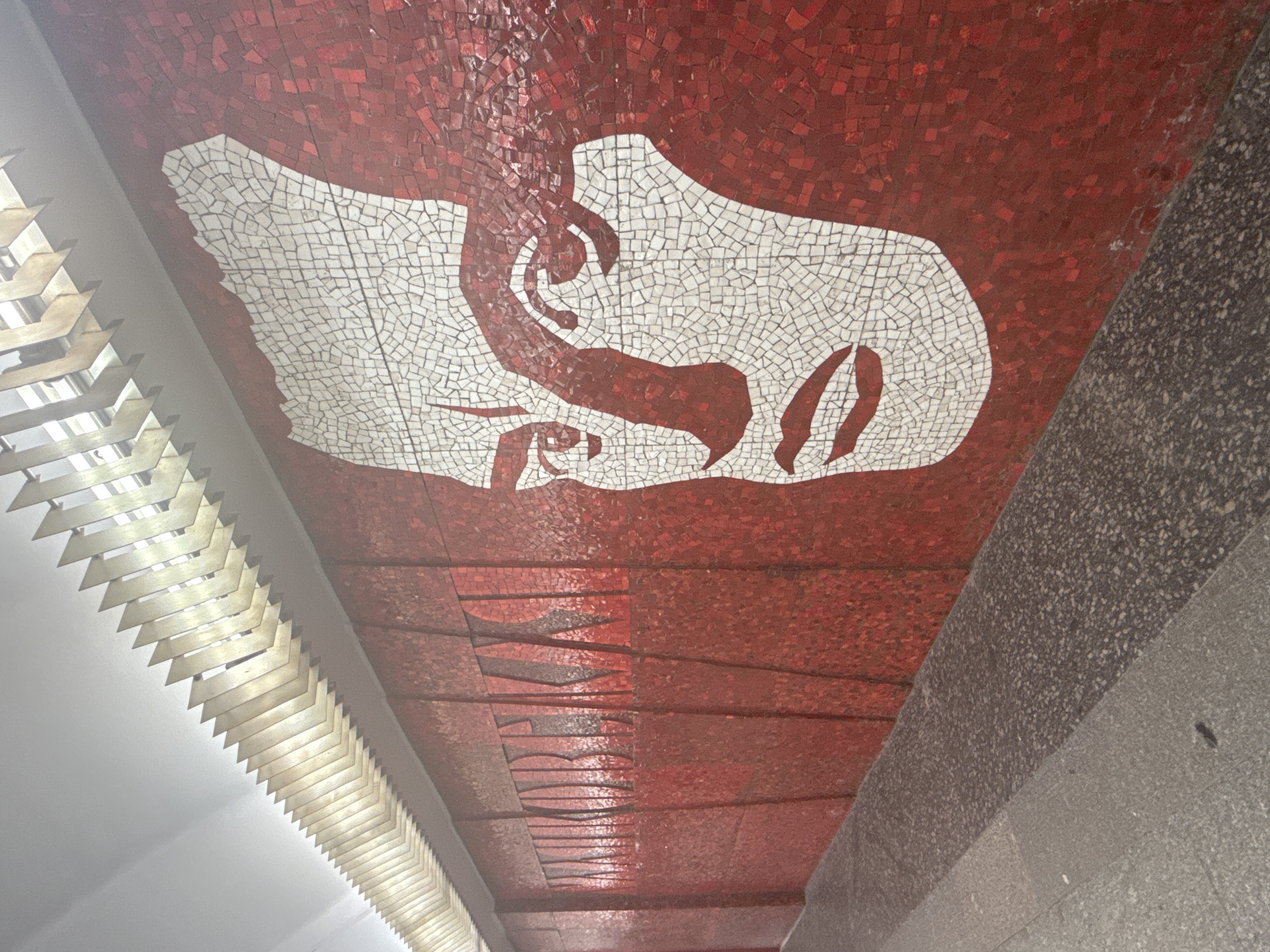 The image depicts a mosaic mural on a wall in a metro station, likely in Saint Petersburg, Russia. The mosaic features a large, stylized white face on a red background. To the left of the face, there are Cyrillic letters also rendered in mosaic, which appear to spell out "МАЯКОВСКИЙ" (Mayakovsky). The ceiling above features a decorative lighting structure with angled metal panels. The floor in the foreground is tiled with a speckled pattern. The overall impression is one of public art in an urban transit environment.