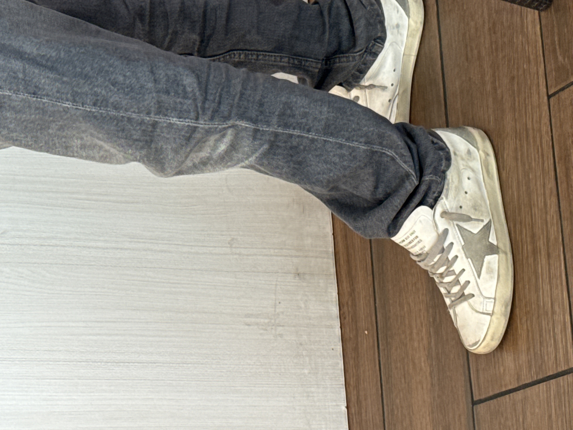 A person is seen wearing gray jeans and white sneakers with a gray star. The sneakers appear worn and dirty. The person is seated. To the left of the person is a surface that appears to be white wood paneling, and to the right is a brown wood surface, possibly a floor or a table. The location is Unknown, Unknown.