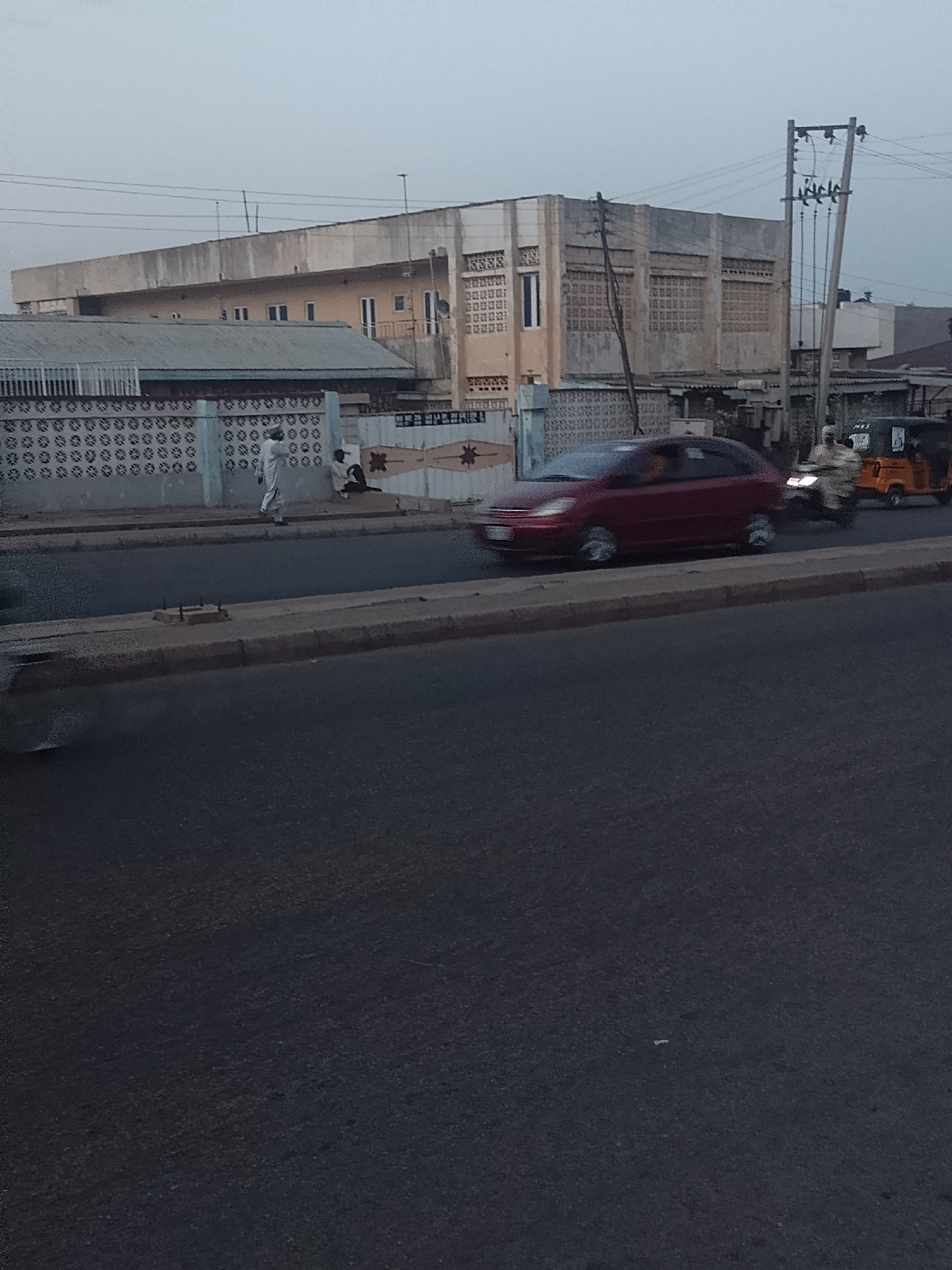 The image depicts a street scene in Pantame, Nigeria. In the background, there is a large, rectangular building with visible architectural details and an adjacent smaller structure with a corrugated roof. A patterned wall or fence runs along the side of the road. A person dressed in white is walking along the sidewalk. A red car is moving along the road. In the distance, there is a motorized tricycle and another motorcycle. Overhead electrical lines and a utility pole are also visible. The foreground is dominated by the asphalt road.