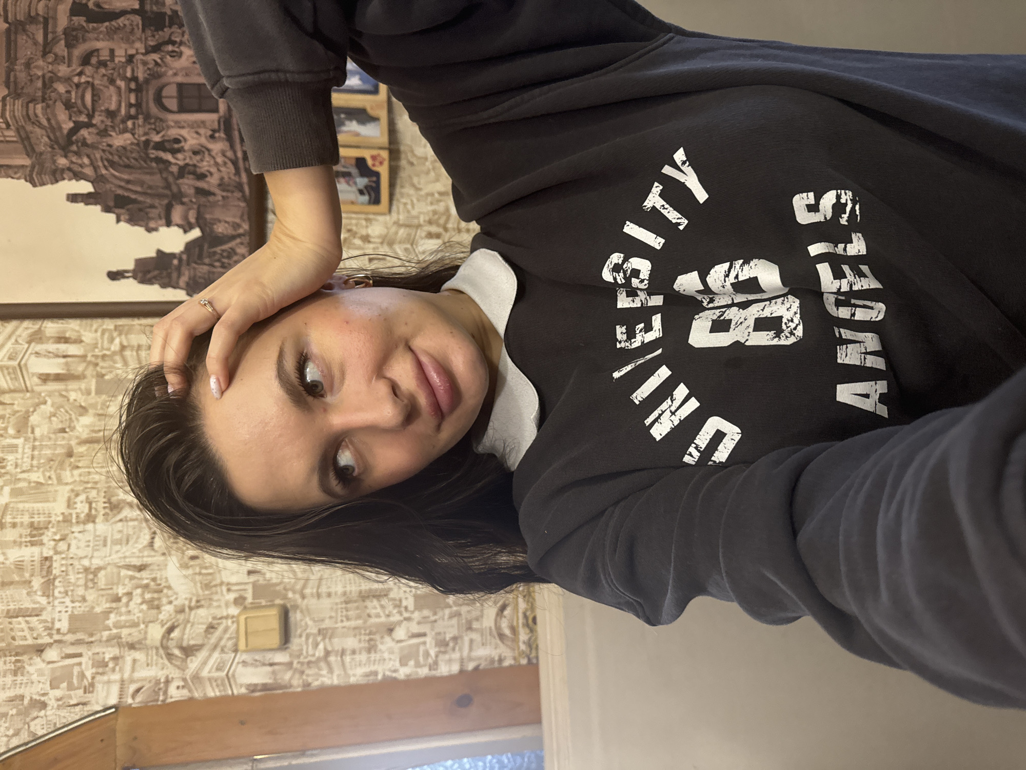 A woman is taking a selfie. She is wearing a dark-colored sweatshirt with white lettering that says "UNIVERSITY 86 ANGELES." She has dark hair, light skin, and is wearing a ring on her left hand. Her right hand is resting on her forehead. The background includes patterned wallpaper, a picture frame, and a window. The location is not discernible from the provided image.