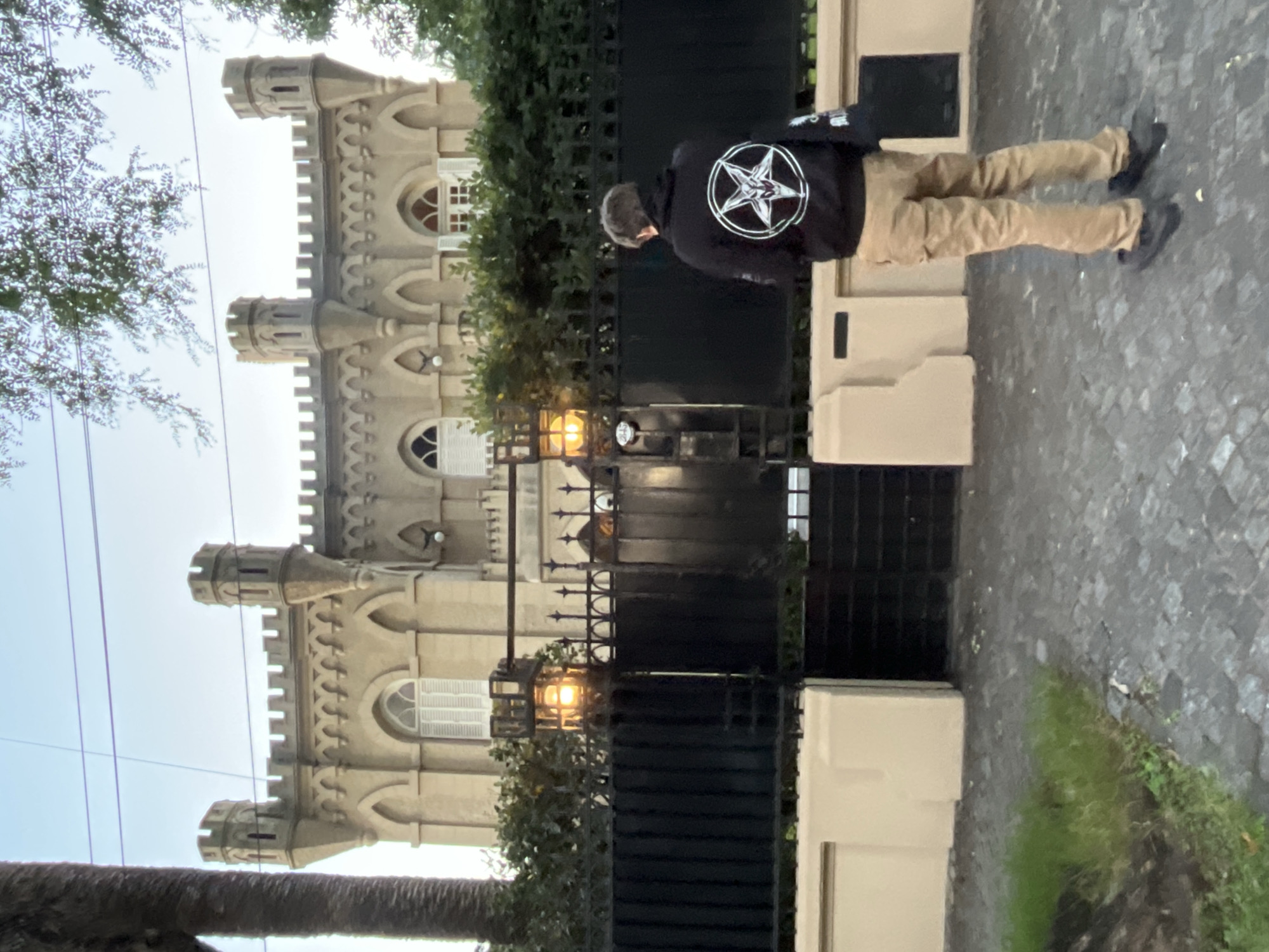 A vertically oriented photograph captures a scene at what appears to be dusk or early morning, given the soft, diffused light and an illuminated street lamp. The image centers on a person standing on a textured paved surface beside an ornate black metal gate, with a distinctive castle-like building dominating the background.
The individual, seen from behind, is wearing a black hooded sweatshirt and light-brown (khaki) pants. A prominent white circular emblem is emblazoned on the back of the hoodie. This emblem features a five-pointed star (pentagram) that contains a stylized goat's head, widely recognized as the Sigil of Baphomet or related occult symbolism. The person has short, dark hair and is wearing dark shoes.
Behind the intricate black metal gate stands a large, historic-looking building with a fortified or castellated architectural style, characterized by multiple crenellated towers, arched windows, and detailed stone work on its facade. Some windows are fitted with white shutters. Green foliage, including trees and bushes, is visible around the building and partially frames the sky. Utility lines stretch across the upper portion of the sky. The ground consists of dark, uneven paving stones, with a small patch of green grass growing alongside a light-colored wall segment.
The setting, identified as Olivos, Argentina, presents an imposing yet somewhat mysterious ambiance due to the historical architecture and the distinct symbol on the person's attire. No other discernible text is visible in the image.