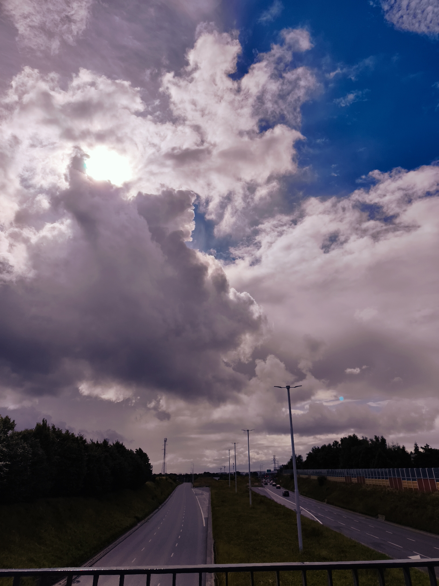 The image captures a view from an elevated position, looking down a multi-lane highway under a dramatic sky. Thick, dark clouds dominate the upper portion of the frame, with patches of bright blue sky visible on the right. The sun is partially obscured by a large, bright cloud formation on the left, casting a diffuse light.
The highway stretches into the distance, with visible lane markings. On the left side of the road, there is a grassy embankment leading up to a dense line of dark green trees. On the right side, the highway is bordered by a sound barrier with sections of yellow, red, and blue panels. Several streetlights are visible along the highway. A few vehicles are driving on the highway. A tall transmission tower can be seen in the distance on the left. The foreground shows the top of a dark metal railing, suggesting the viewpoint is from a bridge or overpass.
While the image does not contain explicit markers of Gdańsk, Poland, the infrastructure and landscape are consistent with the outskirts of a large European city. The presence of a modern highway, sound barriers, and transmission towers, combined with the natural elements like trees and the sky, are commonly found in such environments. Without further context or specific landmarks, it is not possible to definitively pinpoint the location within Gdańsk.