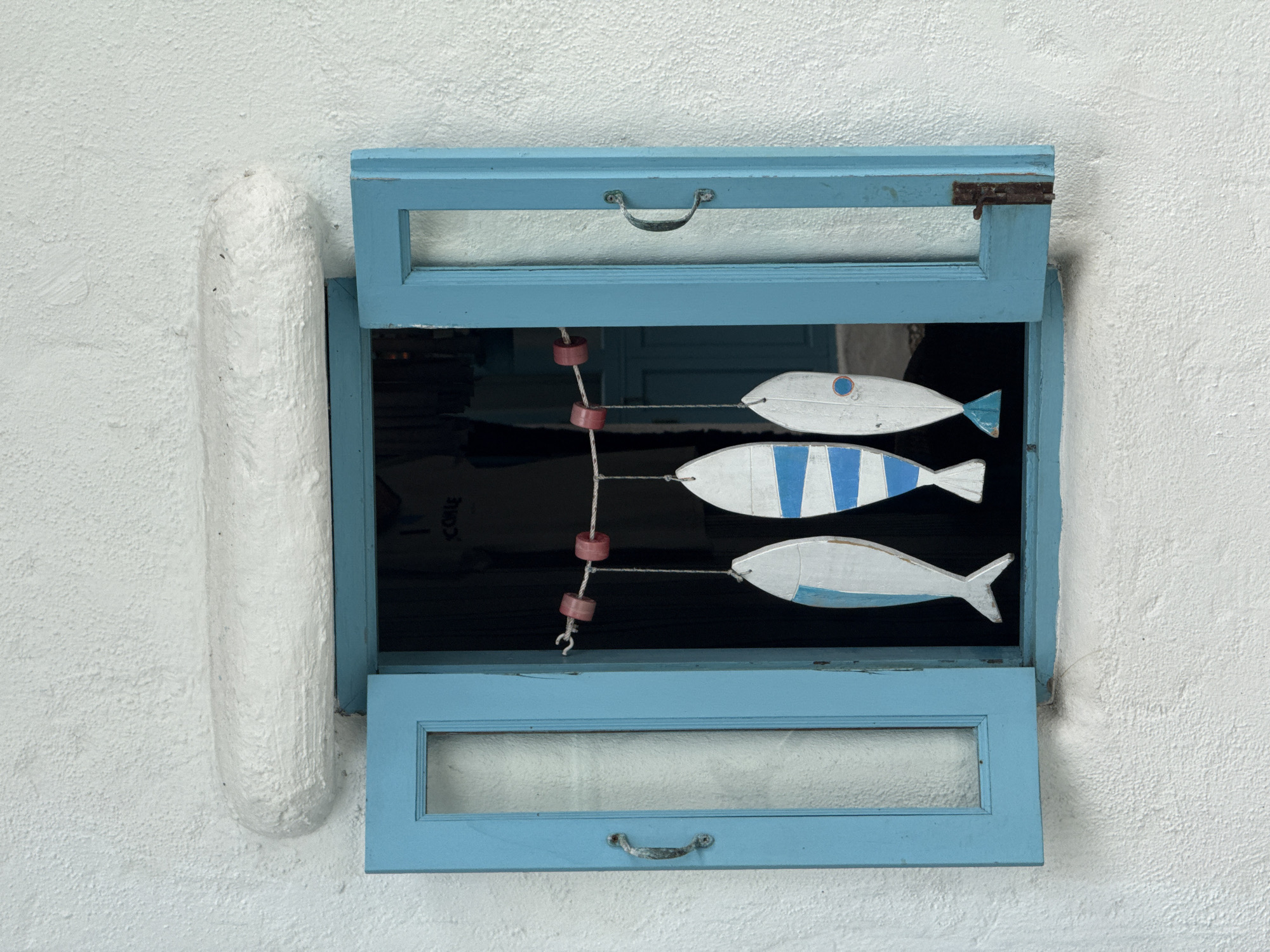An open light blue wooden window with glass panes is set into a rough white stucco wall. Above the window is a thick, rounded white stucco protrusion. Three wooden fish decorations, painted white with blue accents and one with a blue eye, hang from a string with red plastic floats across the window opening. The fish are suspended at different heights. The window frames are slightly weathered, and metal handles are attached to the left and right panes. The interior of the window is dark, showing a hint of clothing with the word "COME" visible. The scene is a close-up shot of the window and wall, with no people visible. The lighting suggests it is daytime, with diffused light casting subtle shadows. There are no specific indicators to pinpoint the exact location within Hua Hin City Municipality, Thailand, beyond the general architectural style.