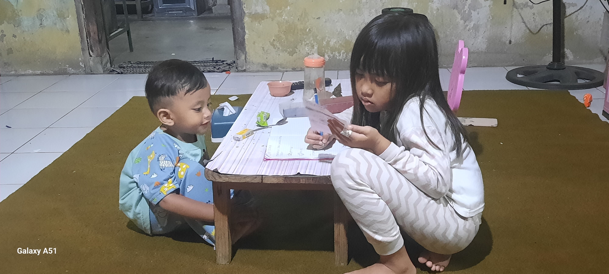Two children, a young boy and an older girl, are sitting on a brown carpet in front of a low wooden table. The boy, wearing a blue pajama set with cartoon animals, is on the left, looking towards the table. The girl, on the right, in a white pajama set with a chevron pattern, is focused on a piece of paper she is holding and appears to be writing or drawing with a pen. The table has a pink and white striped cover. On the table are several items, including a bottle with an orange cap, a small pink bowl, a box of cigarettes, a spoon, and papers. The background shows a wall with peeling paint, a tiled floor, and parts of furniture and a fan. The scene appears to be an indoor living space in Ploso, Indonesia, likely during the evening given the lighting.