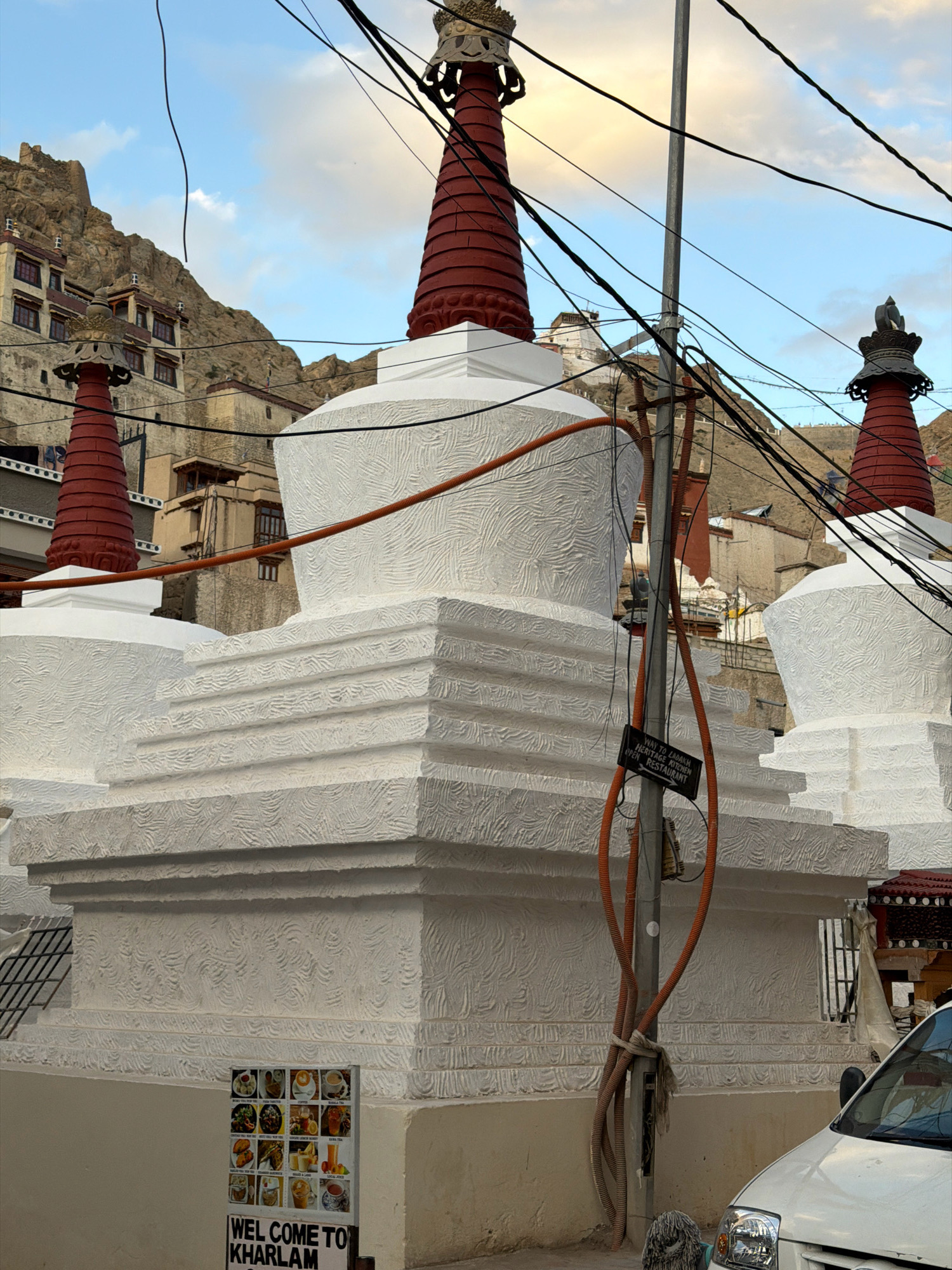 A white stupa with intricate carvings and red spires is in the foreground, with a power pole covered in orange electrical conduits and wires running vertically in front of it. To the left of the stupa, a sign displays a grid of food images and the text "WEL COME TO KH" below. In the background, more stupas and Tibetan-style buildings are nestled against a rocky, mountainous landscape under a cloudy sky. A white car is partially visible in the bottom right corner. This scene is located in Leh, India.