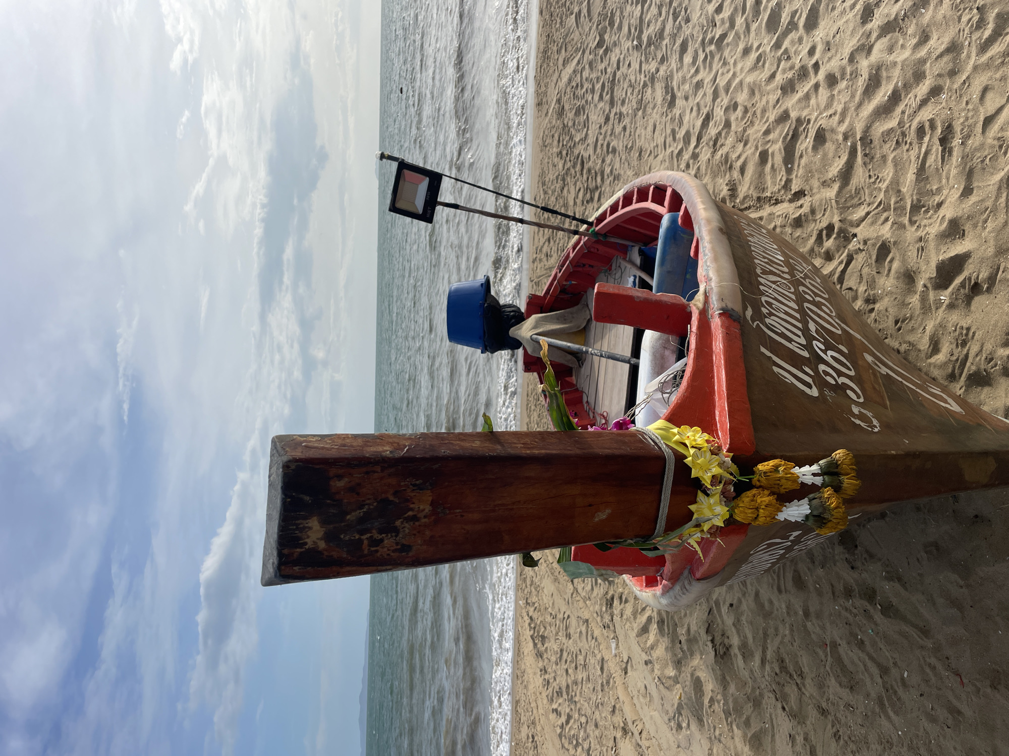 A long-tail boat is beached on a sandy shore in Pattaya City, Thailand, facing the ocean. The boat, painted red and brown, has a prominent wooden pole at its bow, adorned with yellow and white flower garlands. Inside the boat, a blue bucket, a tan cloth, and other equipment are visible. A metal pole with a black rectangular fixture, possibly a light or sign, stands near the stern. The ocean, with gentle waves, stretches to the horizon under a cloudy blue sky. The sand in the foreground is textured with ripples from the wind and tide.