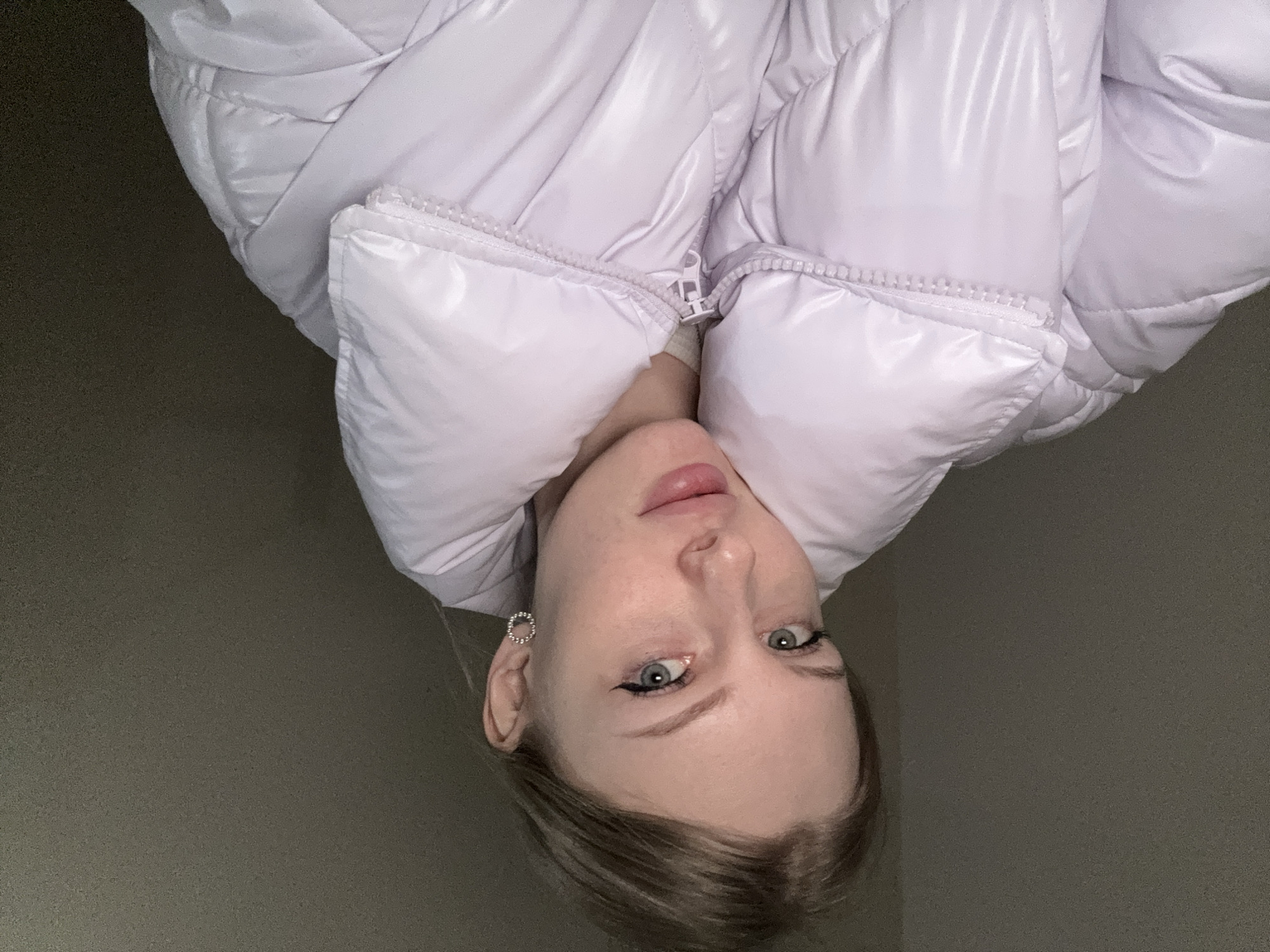 A young adult female is depicted in a close-up, chest-up portrait against a plain, dark gray, textured background, indicating an indoor setting in Perm, Russia. She has light-colored hair, pulled back, and light-blue or green eyes with black winged eyeliner visible on both upper lids. Her lips are slightly parted, revealing a natural pink hue. She wears a light lavender, glossy, quilted puffer jacket with a high collar and a visible white zipper closure. A single circular, beaded or studded earring is visible on her right earlobe. Her head is slightly tilted, and she gazes directly forward, engaging the camera. No other individuals or significant objects are present within the frame.