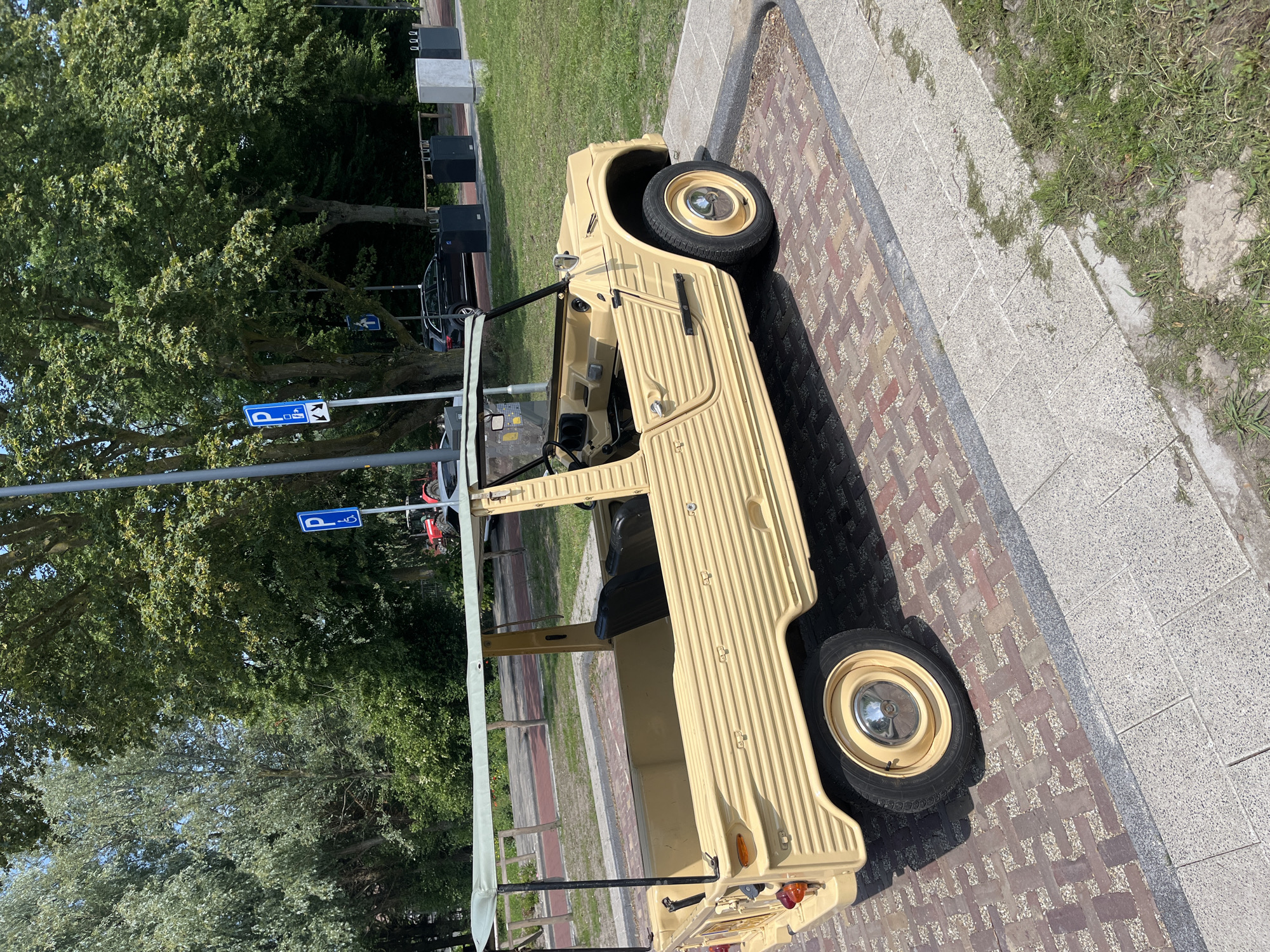 The image shows a vintage, light yellow open-top vehicle parked on a brick-patterned pedestrian walkway. The vehicle has a roll-back canvas roof supported by black poles and a ribbed body design. It is a two-door car with black seats visible inside. The car is positioned at an angle, with its front facing left and slightly away from the camera. In the background, there are trees, grass, and two blue parking signs with the letter "P" and symbols indicating accessibility. A dark-colored SUV is parked further in the background behind the vintage car. The environment appears to be an urban or suburban outdoor setting on a sunny day.