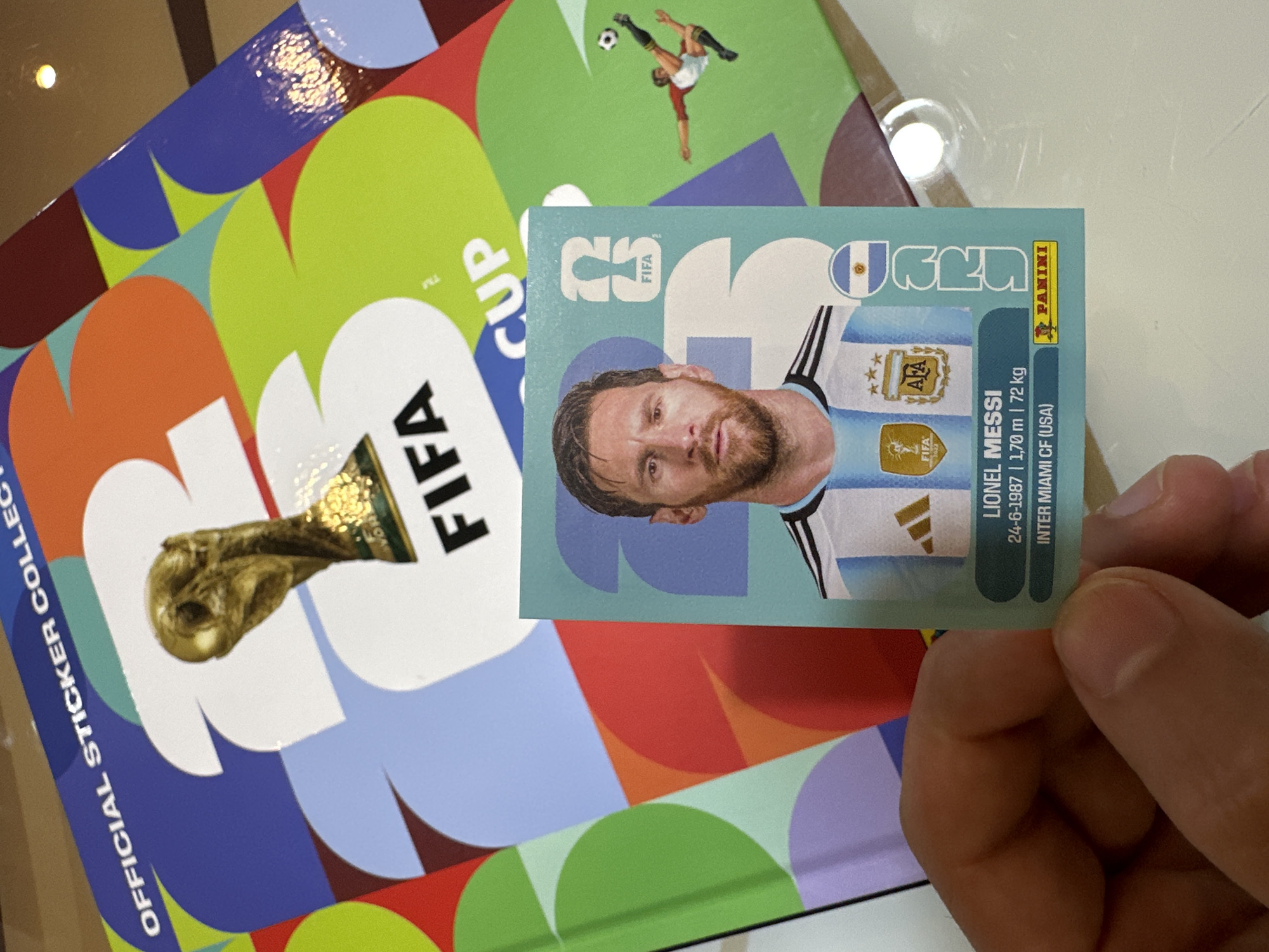 The image shows a close-up of a hand holding a Lionel Messi FIFA World Cup sticker. The sticker features a portrait of Messi in his Argentina kit, along with his name, birthdate, height, weight, and club affiliation (Inter Miami CF). The sticker also includes the FIFA World Cup logo and the Panini logo. In the background, a larger FIFA World Cup sticker album is visible, featuring a golden trophy and the text "OFFICIAL STICKER COLLECTION". The album has a colorful geometric design. There is no indication of the city of Prisovo, Bulgaria, in the image.