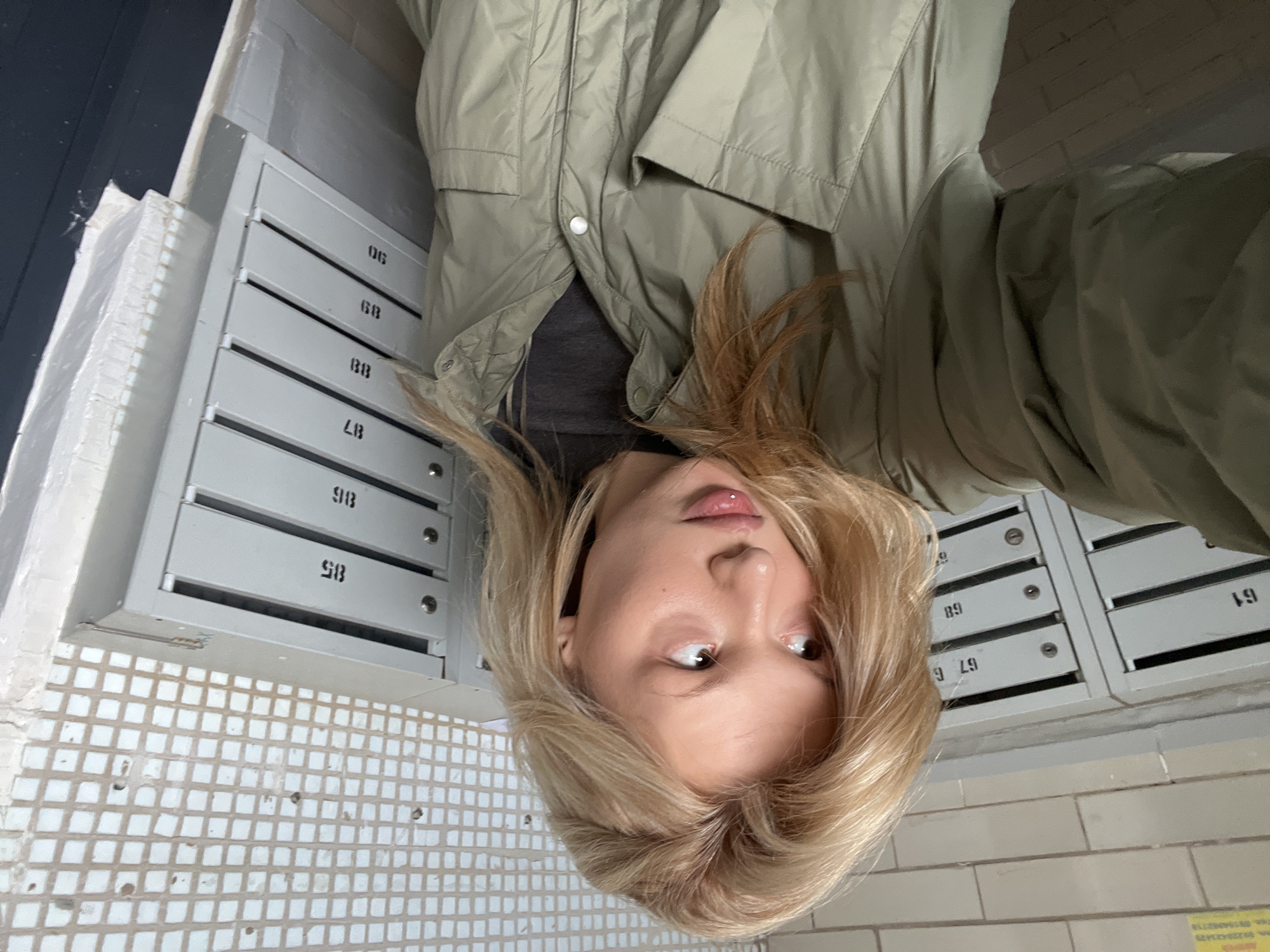 A young female with blonde hair and a lip piercing is depicted in a selfie, looking upwards and to her right. She wears a light olive-green jacket over a dark gray shirt. The setting is an interior hallway of an apartment building, characteristic of residential architecture in Chaikovsky, Russia. Two banks of stacked, grey metal mailboxes line the background. The left bank displays numbers 61, 67, and 68. The right bank displays numbers 85 through 90. The wall behind the left mailboxes is light-colored brick, with a small yellow sign in the upper left corner containing indistinct Russian text. The wall behind the right mailboxes features small, square, light-colored tiles, with some areas of damage. A white-painted wall section with rough textures is visible to the far right. The floor is dark.