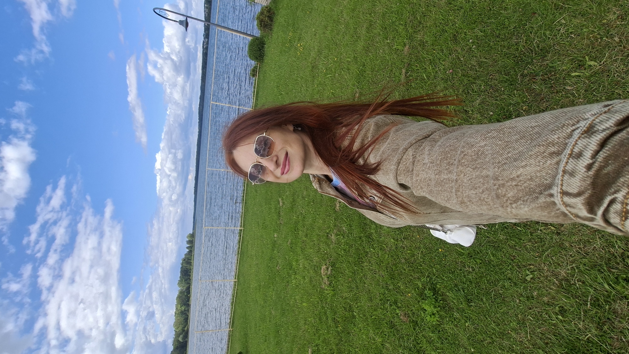 A woman with long red hair and sunglasses is taking a selfie outdoors on a cloudy day. She is wearing a tan jacket over a colorful t-shirt and brown pants. A white purse is attached to her belt. She is standing on a grassy area next to a body of water, with trees and a fence in the background. The scene appears to be a park or waterfront area in Myadzyel, Belarus.
